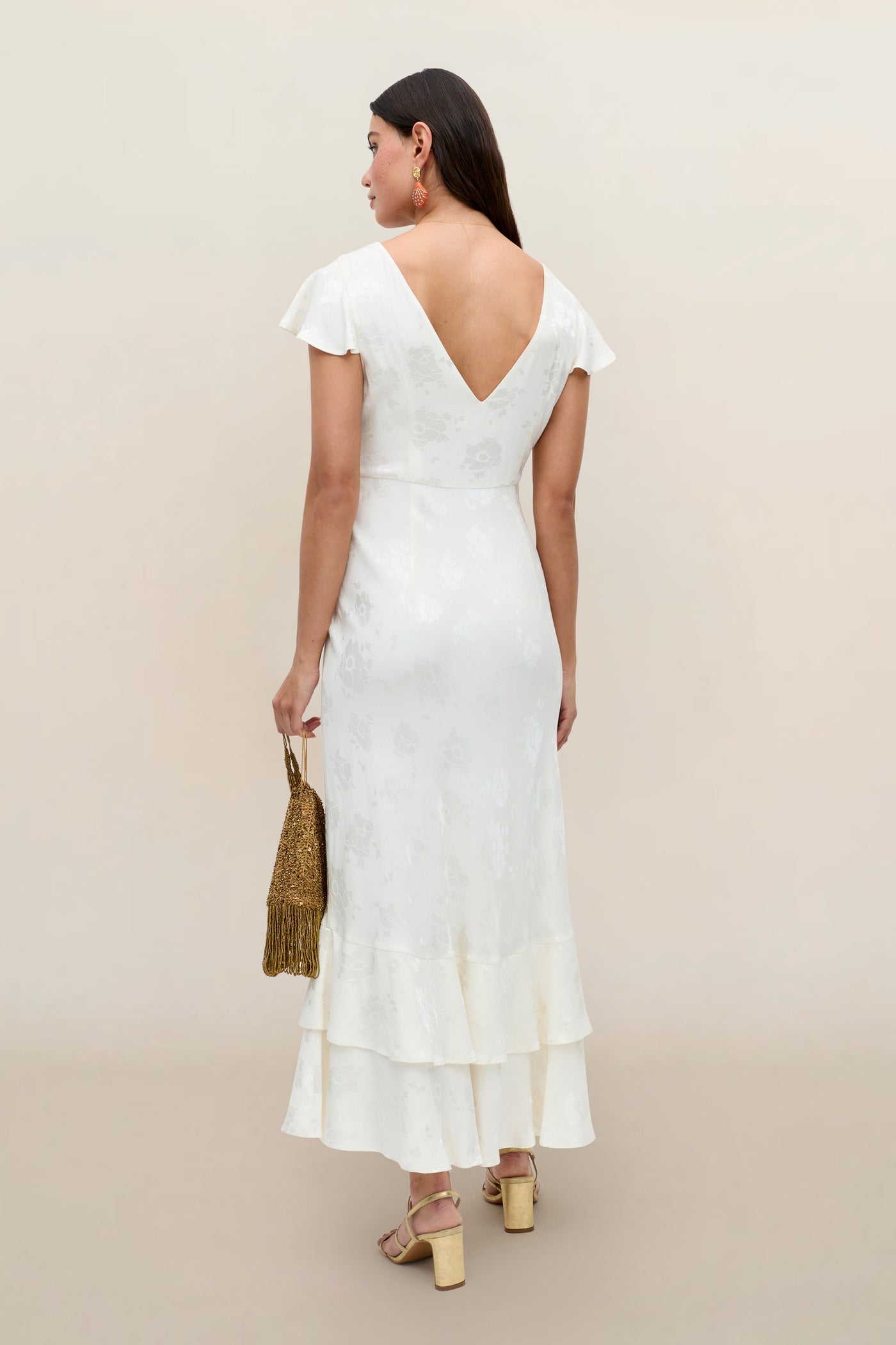 A woman faces away in the Liberty - Poppy ivory jacquard midi dress with cap sleeves, V-back, and ruffled hem. She carries a gold beaded handbag and wears chunky heeled sandals against a light beige background.