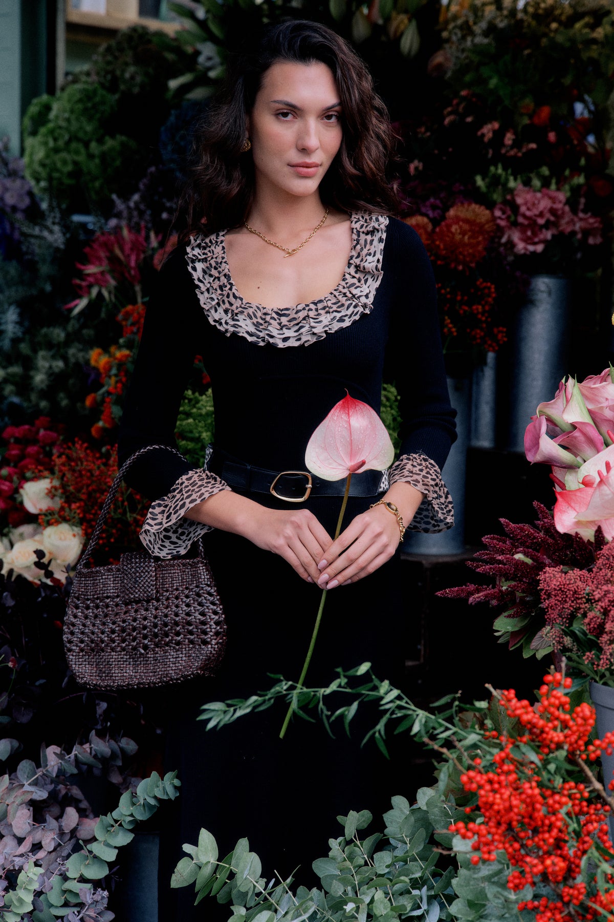 A woman in the Mika - Antelope Spot Black scoop neck dress with ruffled leopard trim stands among colorful flowers, holding an anthurium. She has wavy brown hair, a belt, gold necklace, and a woven handbag.