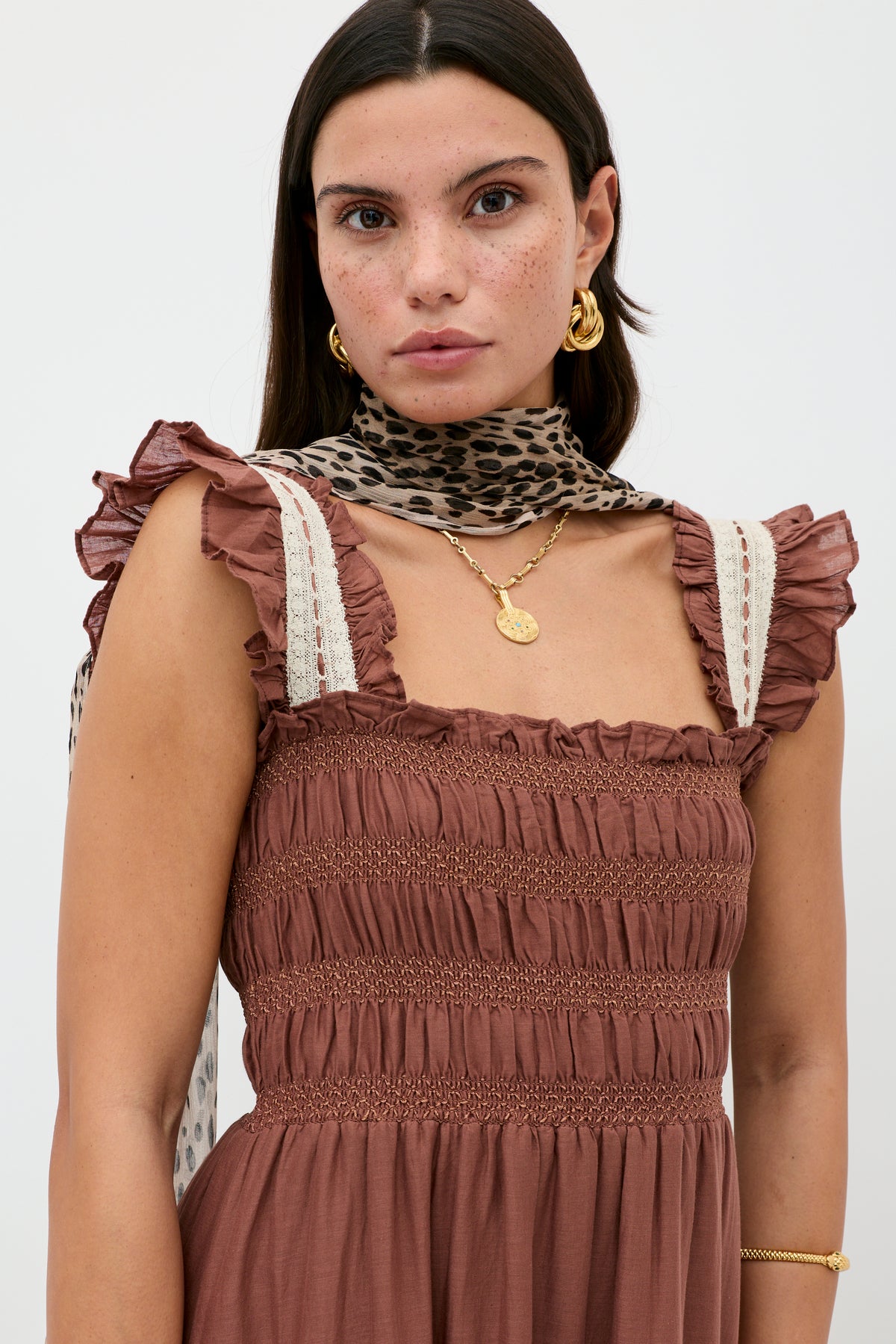 A woman with long dark hair and freckles wears the vintage-inspired Aubrie - Brown dress featuring lace crochet midi details, paired with gold jewelry and a leopard-print scarf, posing against a plain light background.