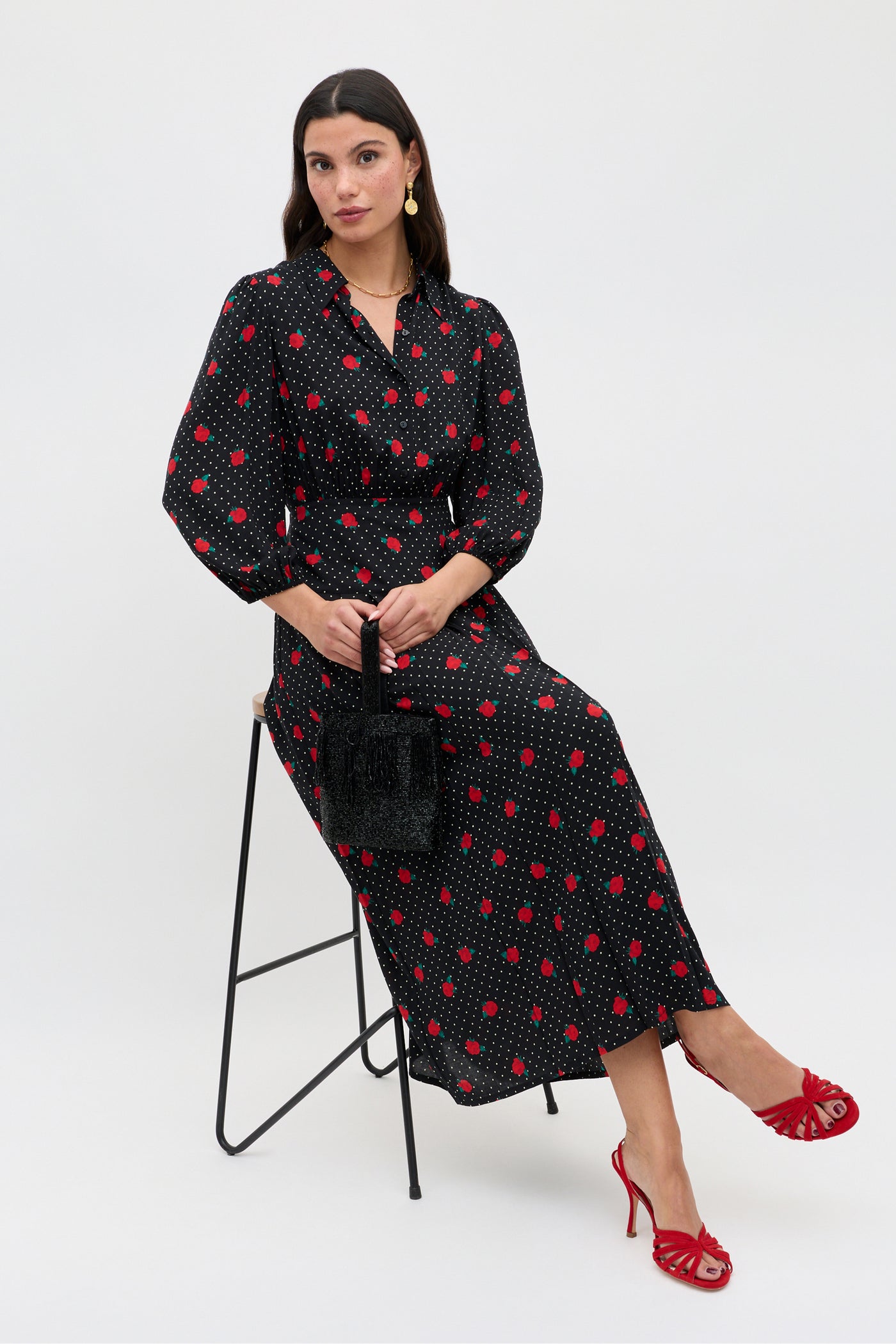 A woman in a long black dress with red polka dots sits on a modern stool, holding a small black bag. She wears gold earrings and glamorous Mavi - Red sandals, posing against a plain white background with dark hair.