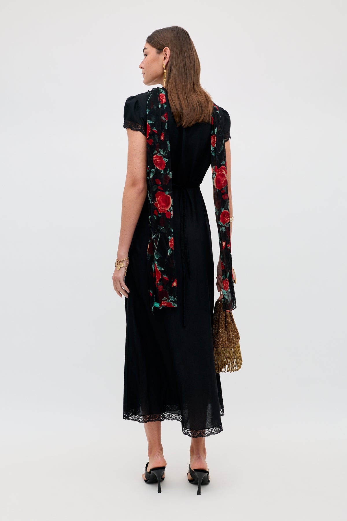 A woman stands facing away, wearing the Julien - Claudia Rose Black short-sleeve dress featuring a lace hem. She accessorizes with a long red floral hand-painted vintage scarf, gold fringed handbag, black heels, earrings, and a bracelet. Her hair is down, and the look is elevated by the fusion of timeless elegance and artistic detail offered by the scarf.