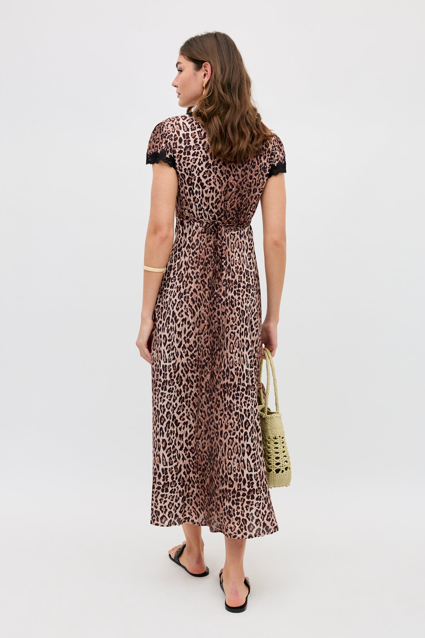 A woman with wavy brown hair stands facing away in the Clarice - Brown Leopard dress, featuring black lace-trimmed sleeves. She pairs it with black sandals, a gold bracelet, and a woven straw bag against a plain white background.