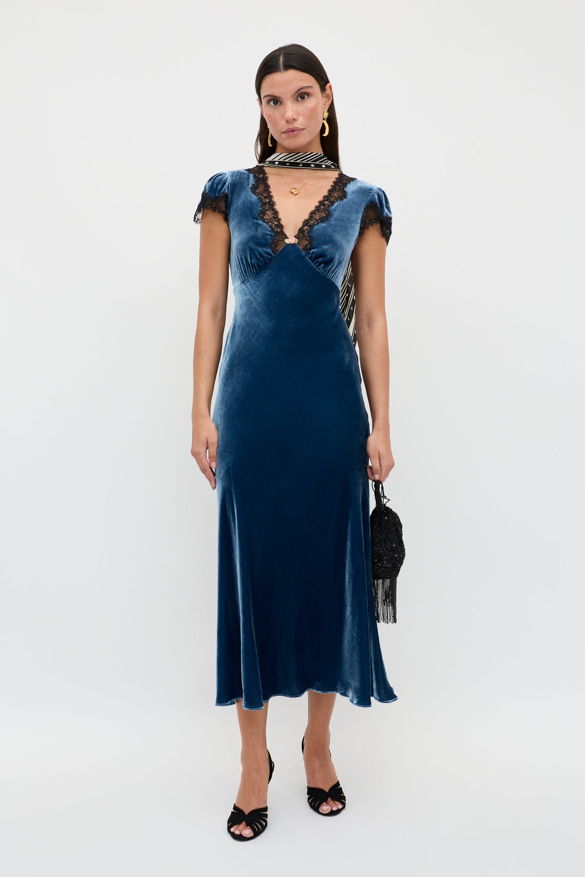 A woman stands against a plain background wearing the Clarice - Steel Blue velvet midi dress with black lace trim and short sleeves, paired with black heels and a small black handbag. She has straight dark hair and minimal jewelry.