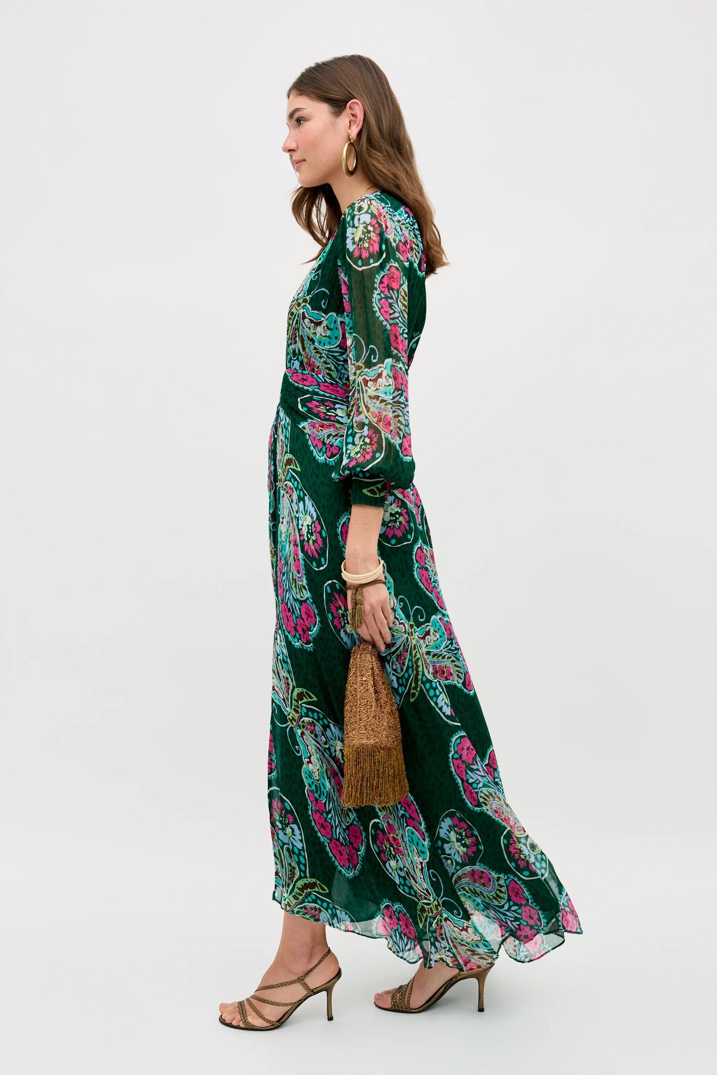 Wearing the Meera - Butterfly Bloom Green dress, a woman walks in profile with a brown textured handbag, strappy heeled sandals, and gold hoop earrings, looking relaxed against a plain light background.