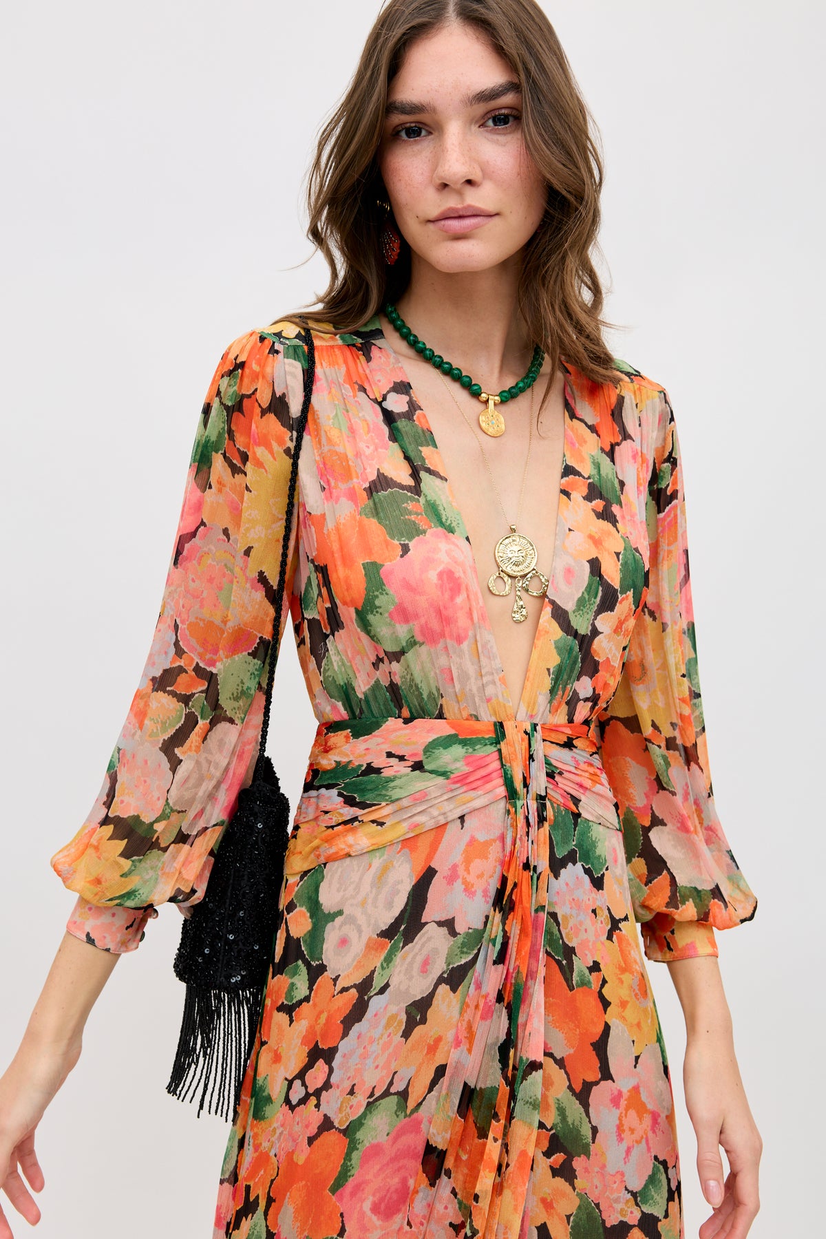 A woman in the Meera - Chelsea Floral Black dress, featuring vibrant florals, a deep V-neck, and long sleeves, stands facing the camera with layered necklaces, loose wavy hair, and a black fringed handbag on her shoulder.