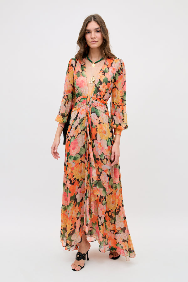 A woman stands against a plain background wearing the Meera - Chelsea Floral Black dress, featuring a long, flowy silhouette with orange, pink, and green patterns, a deep V-neckline, gathered waist, loose brown hair, sandals, and a black shoulder bag.