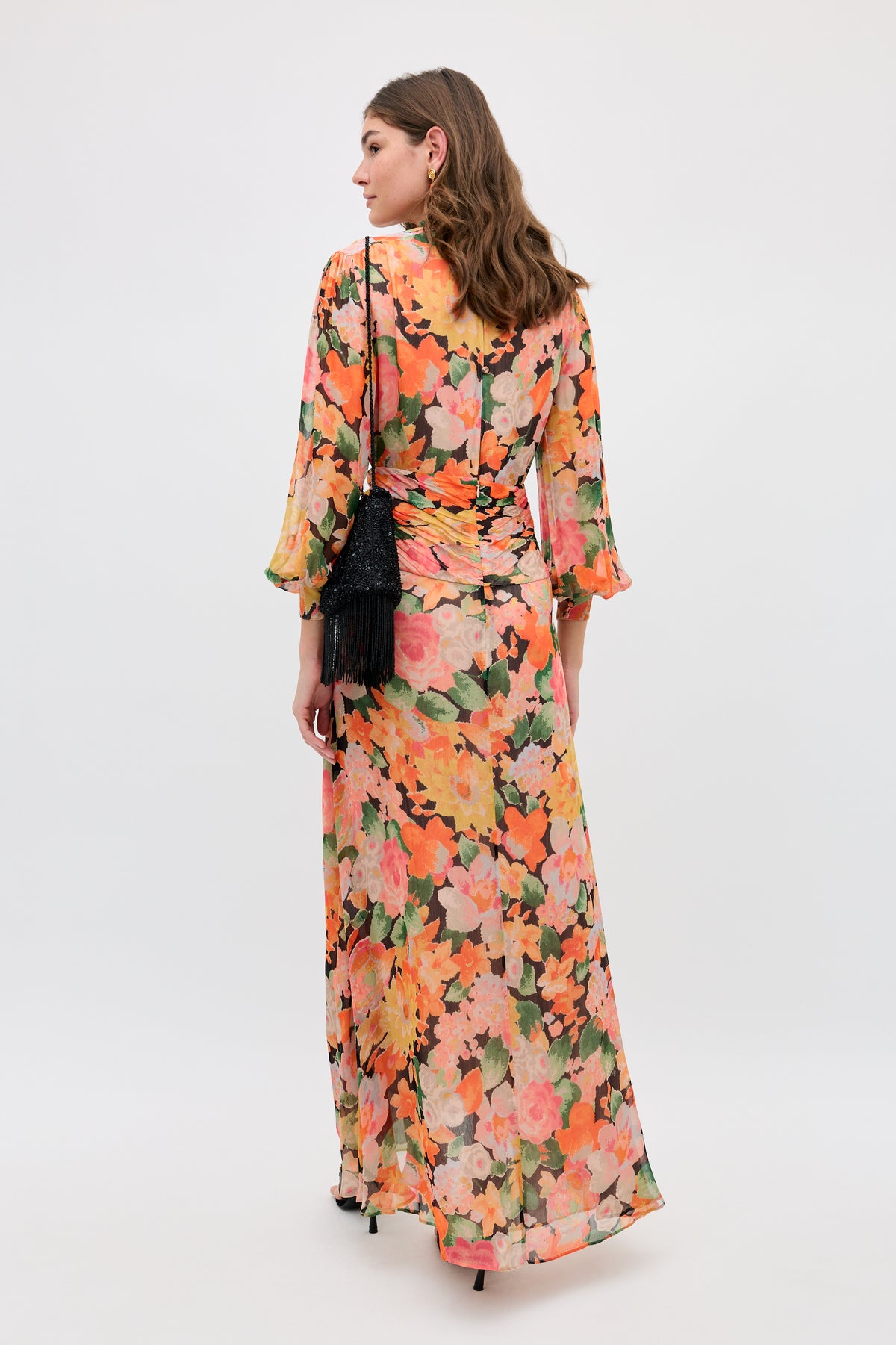 A woman stands facing away, wearing the Meera - Chelsea Floral Black long dress with an orange, pink, and green floral print. She carries a small black fringed purse. The backdrop is plain and light-colored.
