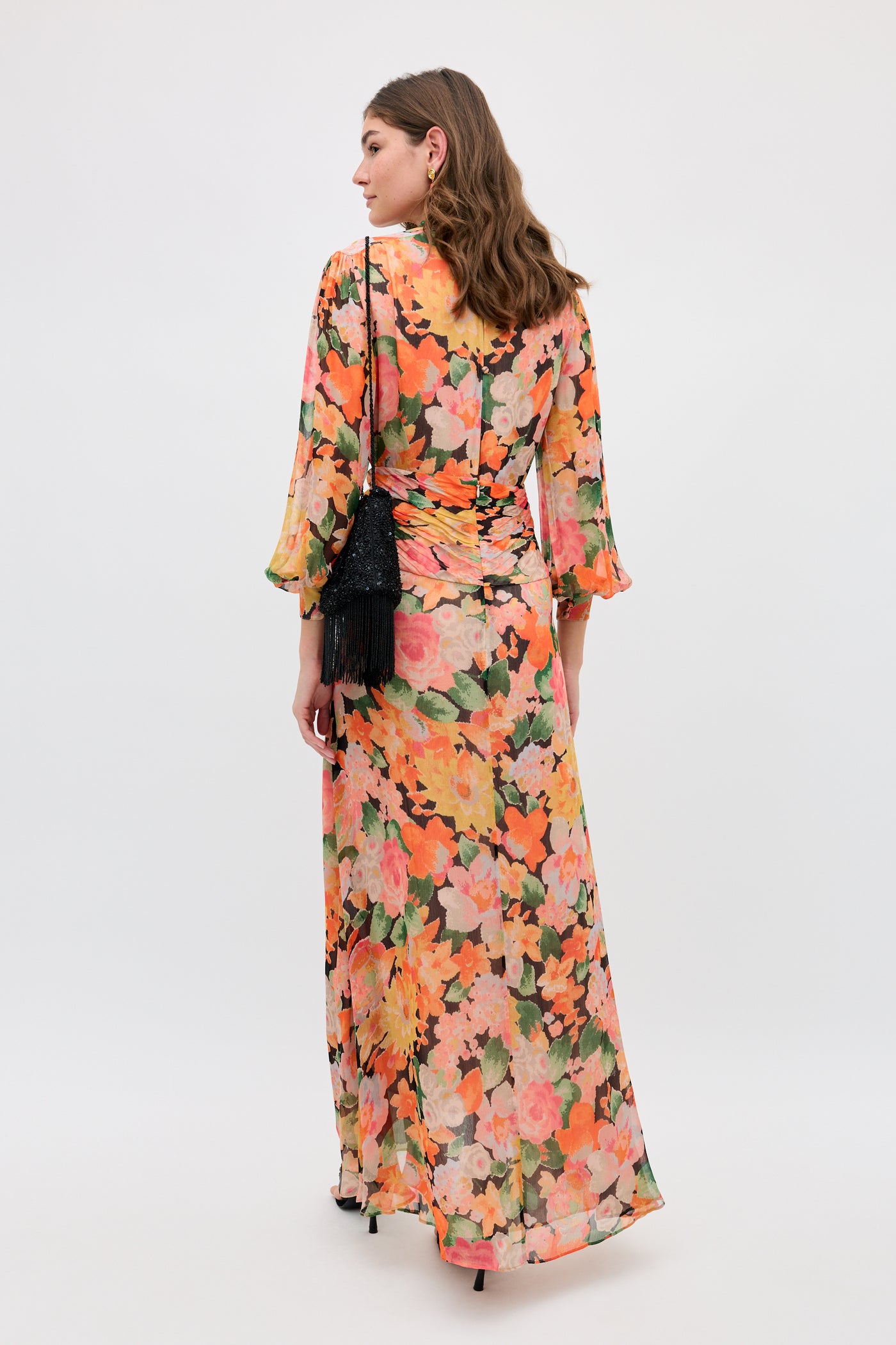 A woman stands facing away, wearing the Meera - Chelsea Floral Black long dress with an orange, pink, and green floral print. She carries a small black fringed purse. The backdrop is plain and light-colored.