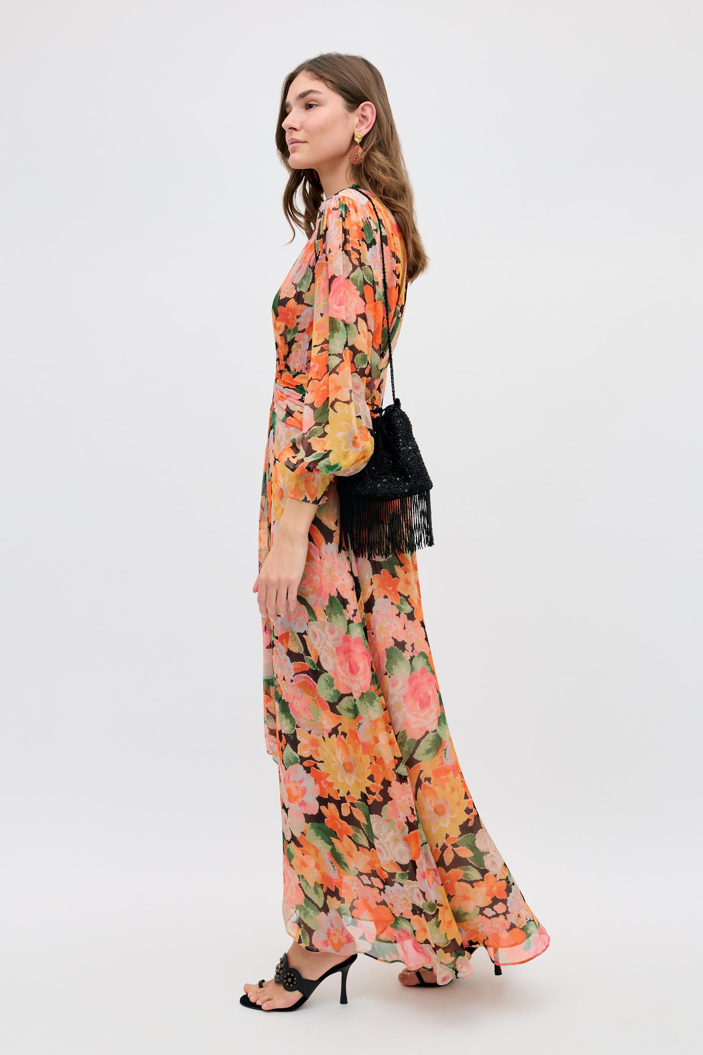 A woman wears the Meera - Chelsea Floral Black long dress with orange, pink, and green patterns. She stands sideways, holding a black fringe purse and wearing black heeled sandals against a plain white background.