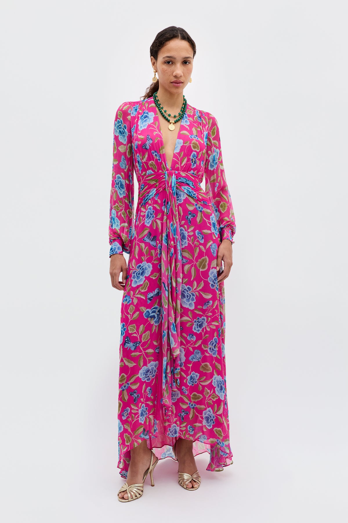 A woman stands against a plain white background, wearing the Meera - Butterfly Floral Magenta dress with long sleeves and a deep V-neckline, paired with a green beaded necklace and gold sandals. She is looking straight ahead.