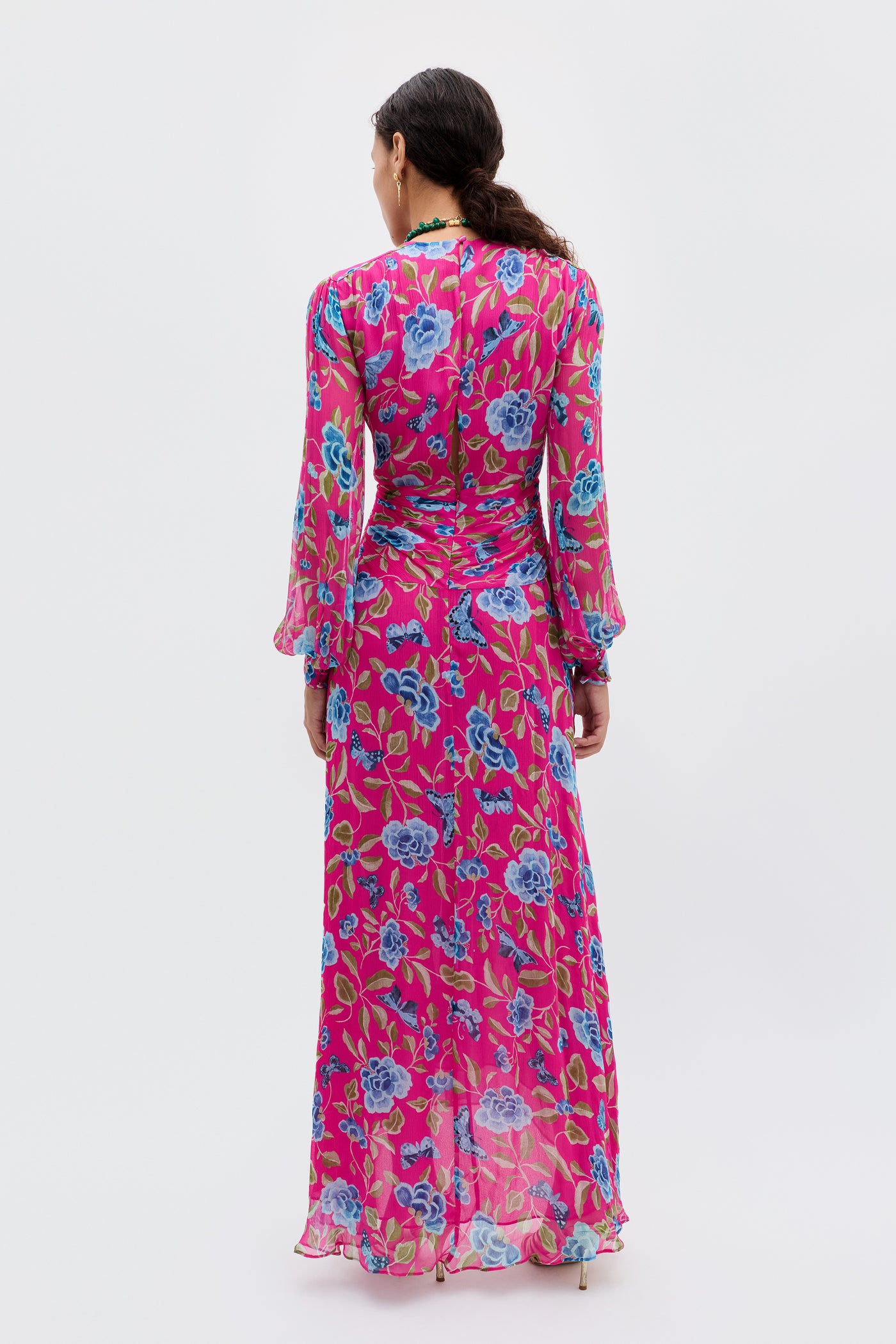 A woman stands with her back turned in the Meera - Butterfly Floral Magenta dress, a long-sleeved, floor-length pink chiffon with blue and green florals. Her low ponytail and the white background highlight this perfect wedding guest look.
