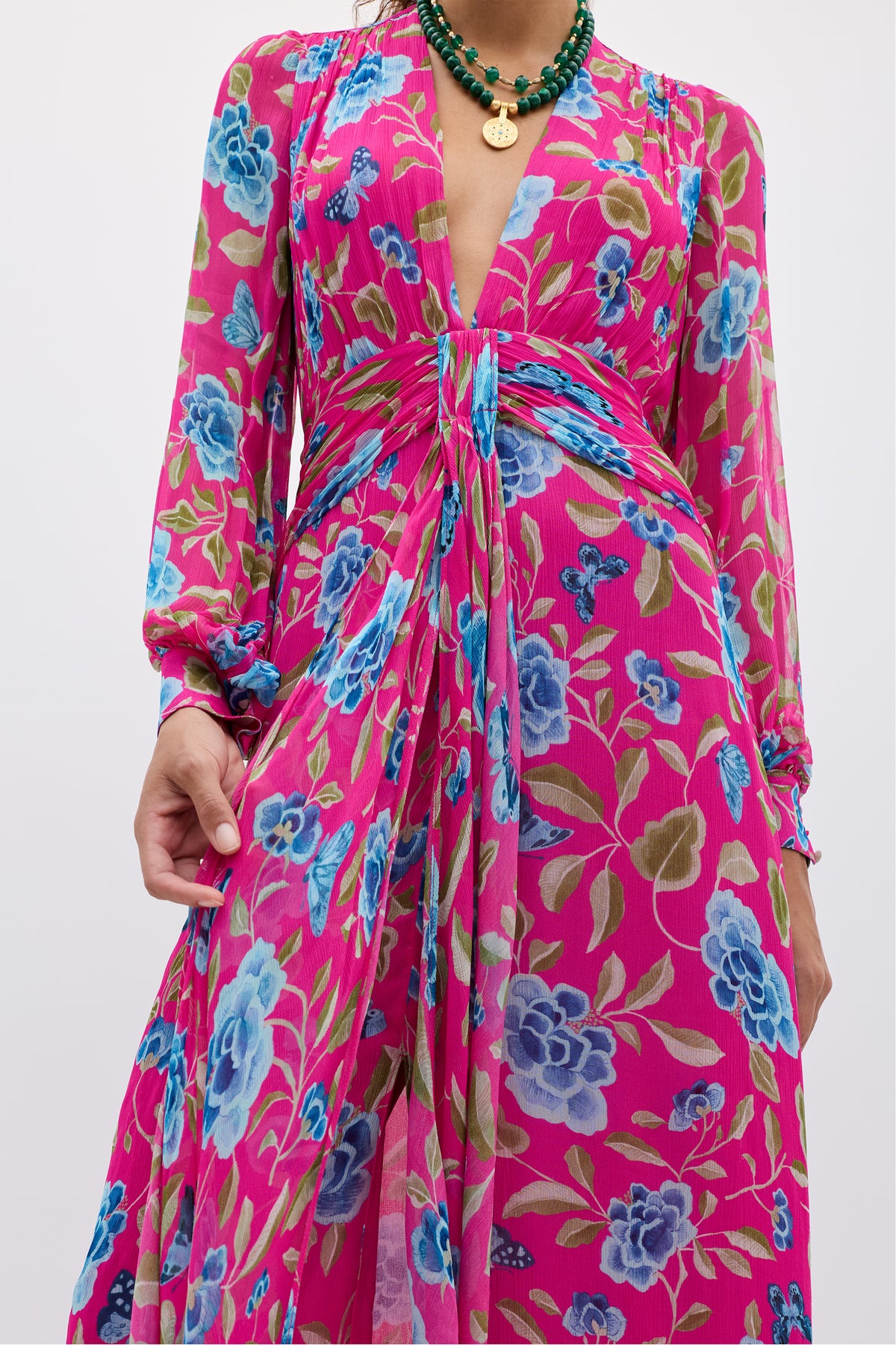 Wearing the Meera - Butterfly Floral Magenta dress, a vibrant pink long-sleeve chiffon piece with blue floral patterns and a deep V-neckline, paired with layered green beaded necklaces and a round gold pendant.