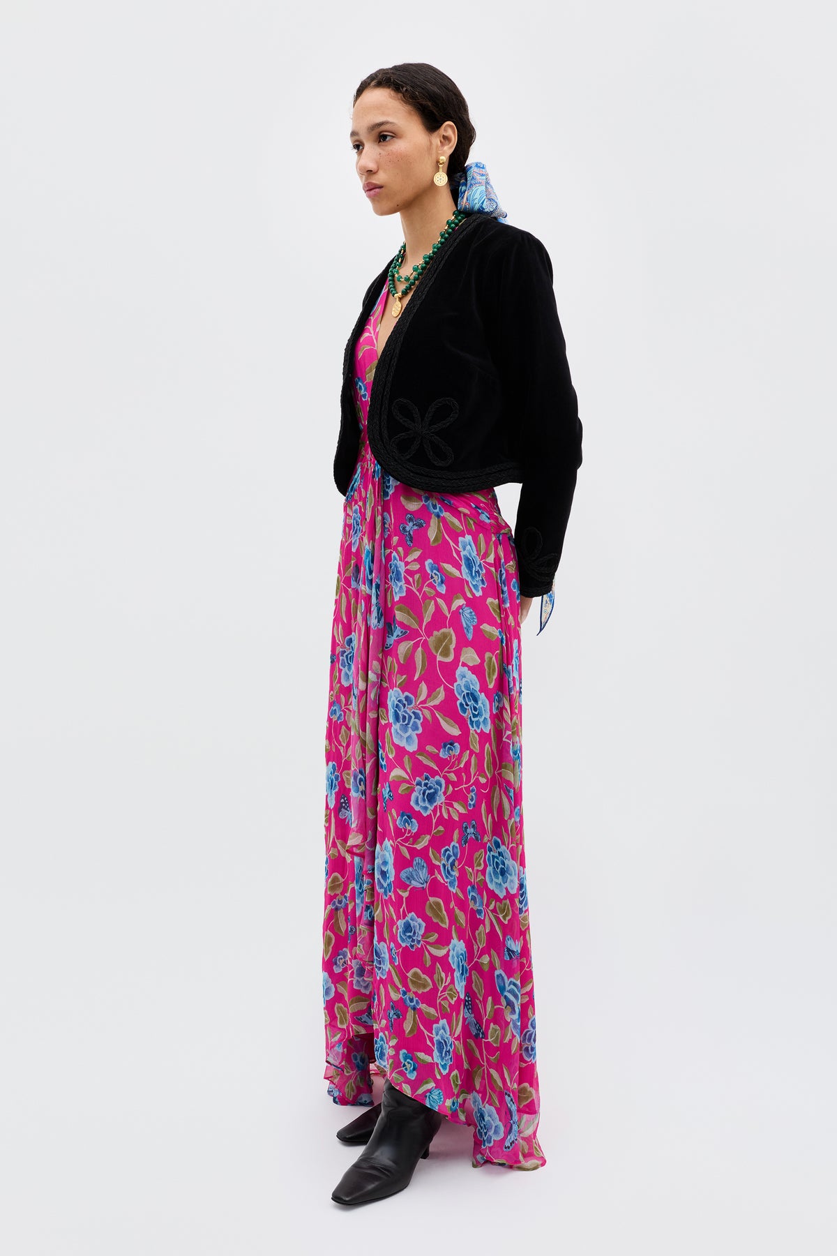 A woman stands in profile wearing the Meera - Butterfly Floral Magenta dress, paired with a black cropped jacket, black boots, a blue hair scarf, and green jewelry, set against a plain white background.