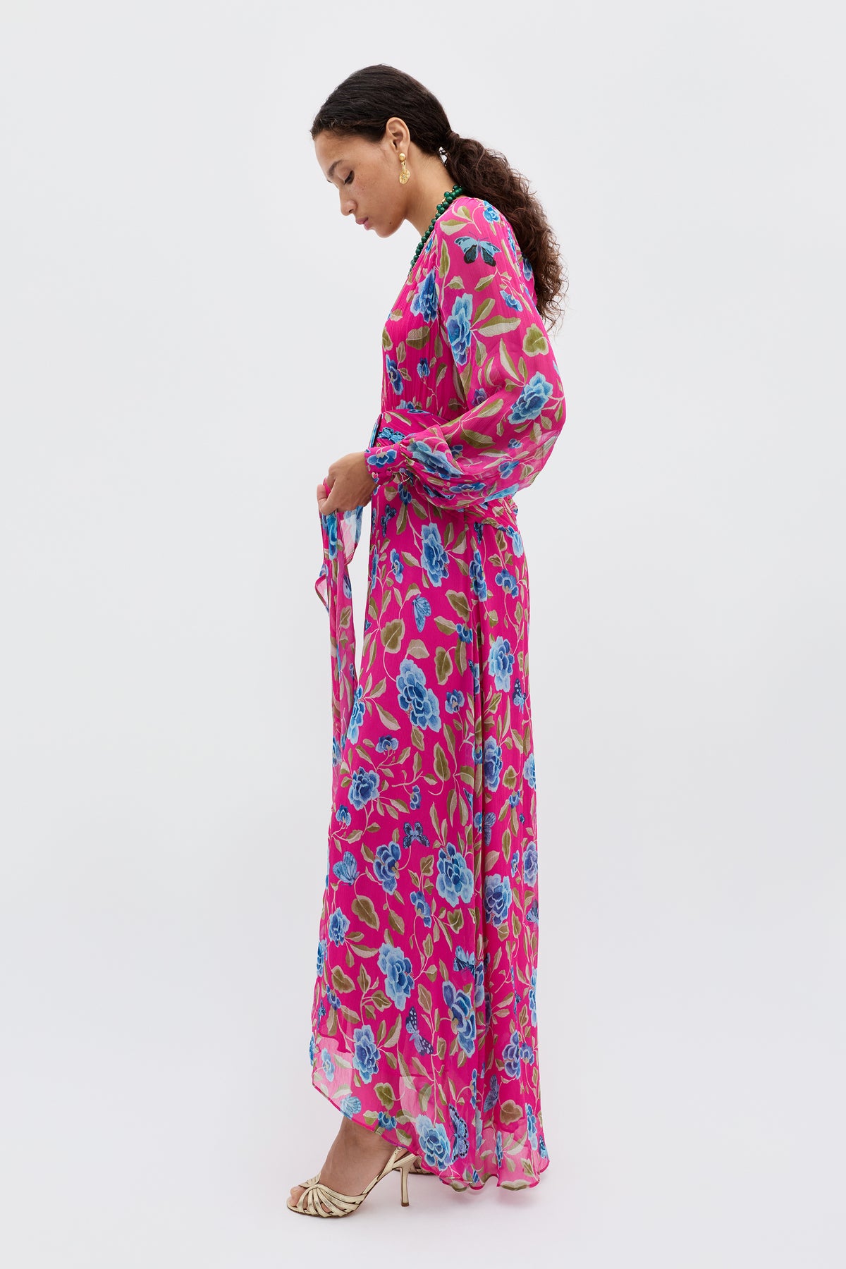A woman stands in profile, looking down, wearing the Meera - Butterfly Floral Magenta dress with curly hair tied back and gold high-heeled sandals against a plain white background.