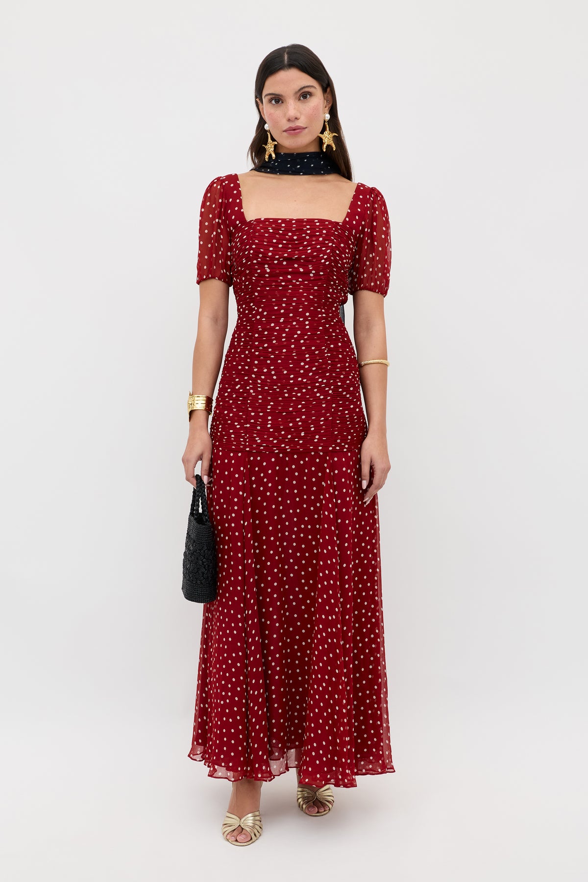 A woman in the Leyane - Layla Dot Red ankle-length, short-sleeve red dress with white polka dots stands against a plain background, styled with gold jewelry, star earrings, a black scarf and bag, and gold sandals.