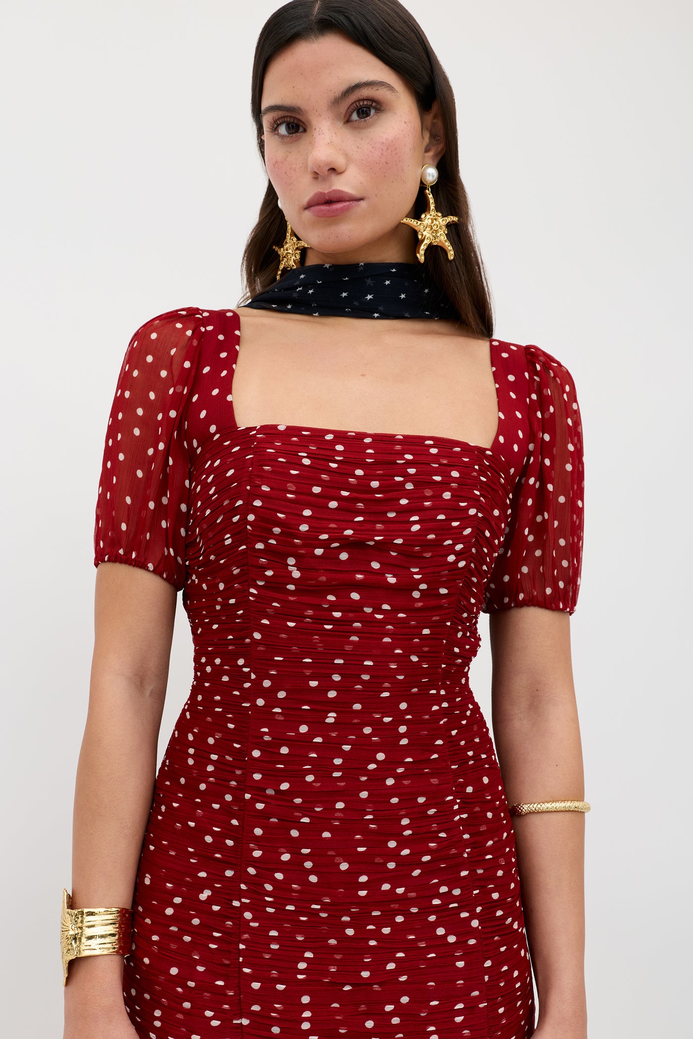 A woman wears the Leyane – Layla Dot Red dress, a red, short-sleeved polka-dot outfit with a square neckline. She accessorizes with gold star earrings, a wide gold bracelet, a gold bangle, and a black scarf with white dots.