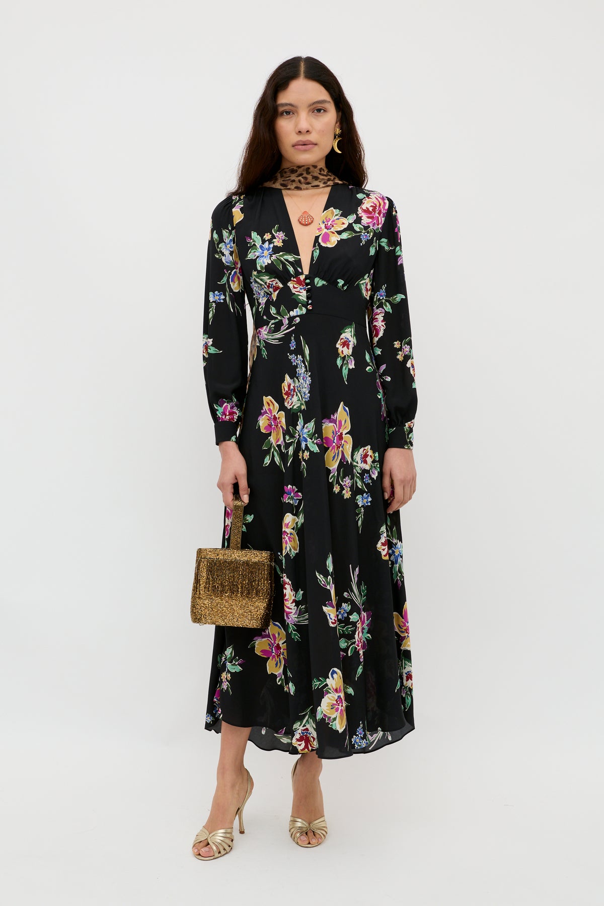 A woman with long, wavy brown hair and a neutral expression stands against a plain background wearing the Trula - Vintage Bloom Black dress—a signature RIXO style featuring long sleeves, black fabric, and delicate floral motifs. She completes her look with gold open-toe heels, a gold handbag, and a black choker necklace.
