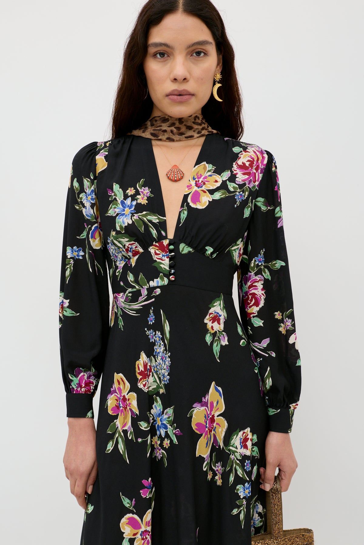 A woman stands against a plain white background wearing the Trula - Vintage Bloom Black, a long-sleeve RIXO maxi dress featuring elegant floral motifs and a deep V-neck. She accessorizes with a leopard print neck scarf, shell necklace, gold hoop earrings, and carries a woven bag.
