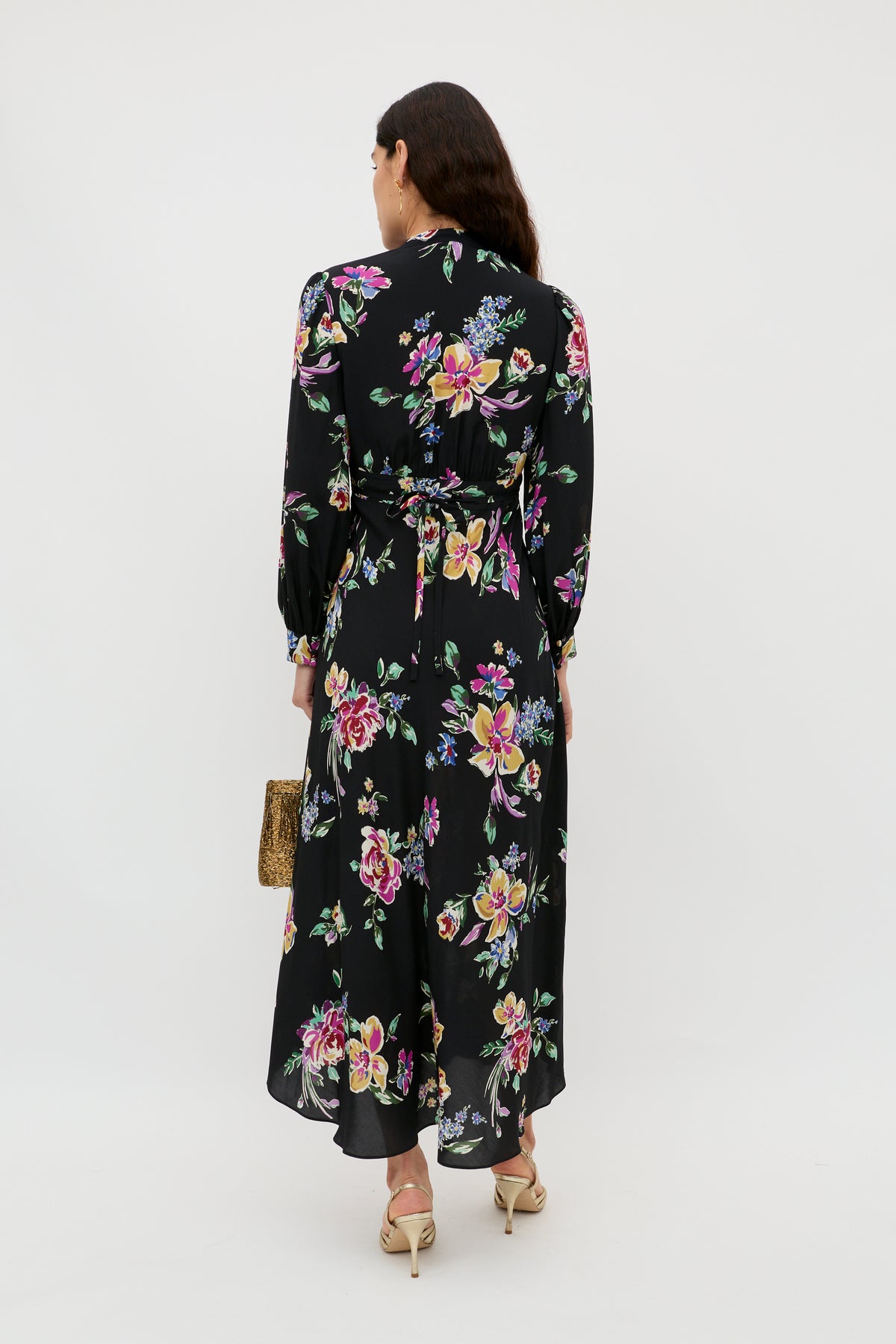 A woman stands facing away, wearing the Trula - Vintage Bloom Black maxi dress by RIXO, featuring vibrant pink, yellow, and green floral motifs. She holds a textured clutch and wears beige heels.