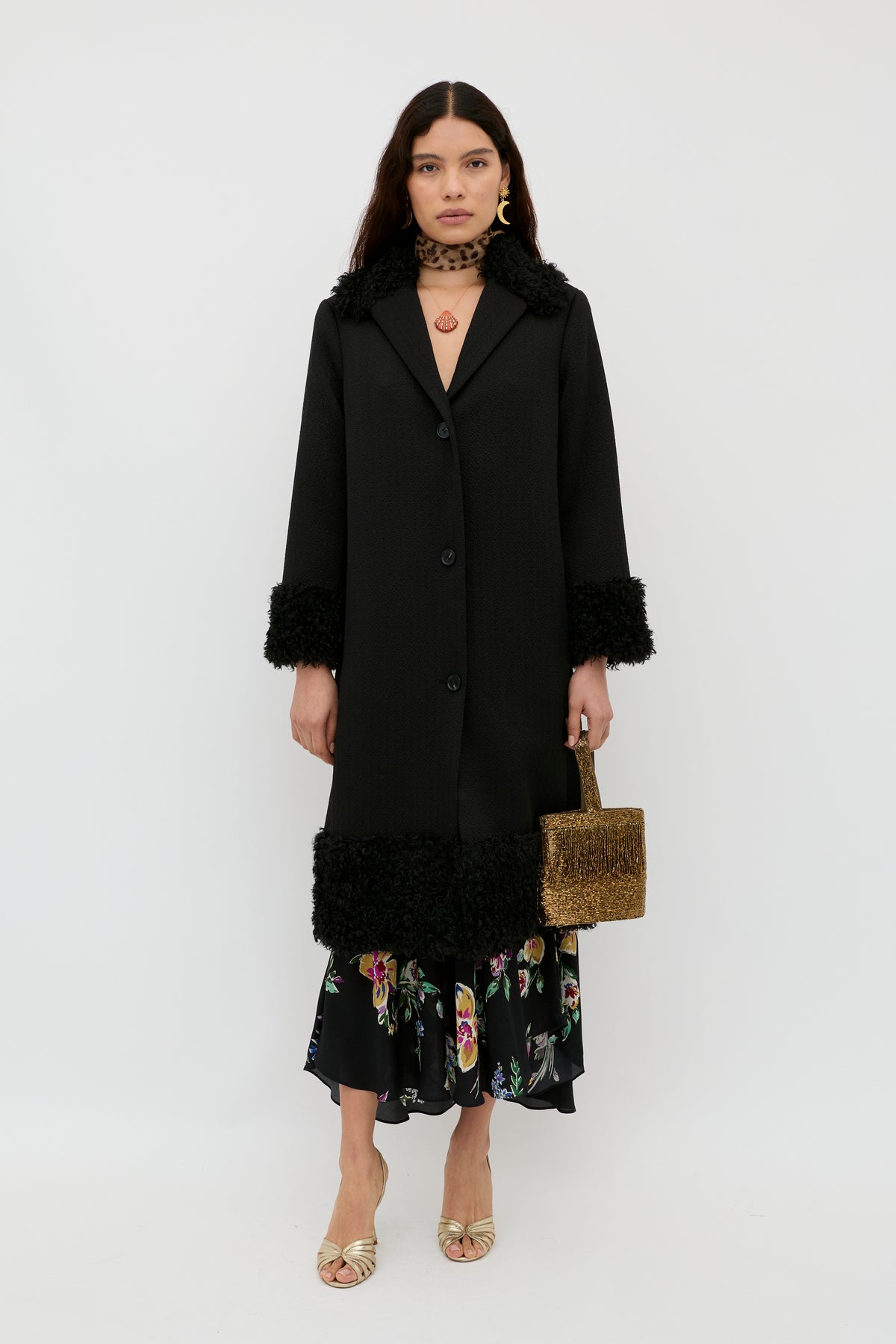 Wearing a black coat with textured cuffs and hem, gold sandals, gold hoop earrings, a brown choker, and carrying a woven handbag, the woman showcases the RIXO Trula - Vintage Bloom Black maxi dress adorned with delicate floral motifs as she stands against a plain white background.