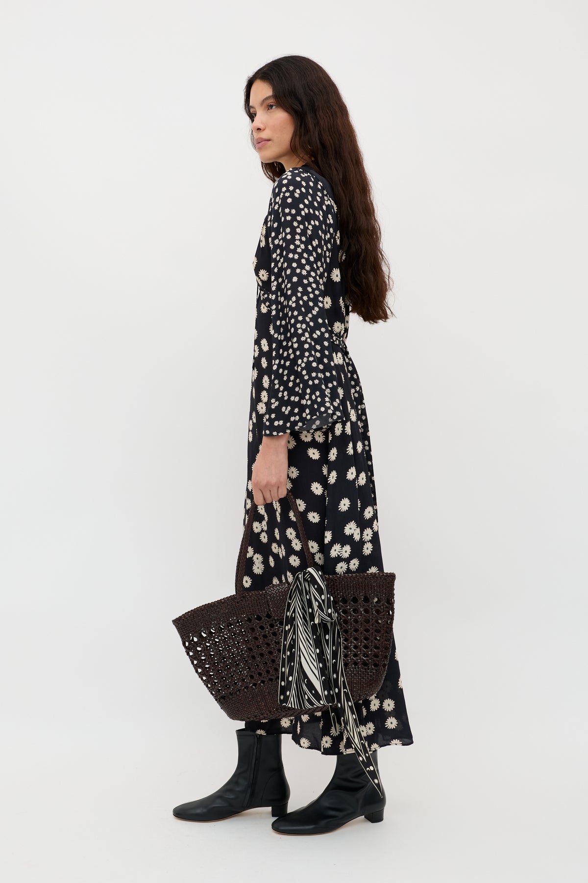 A woman with long dark hair stands in profile against a white background, wearing the Anela - Daisy Mix Black dress, black boots, and holding a large woven bag with a striped scarf tied to it.