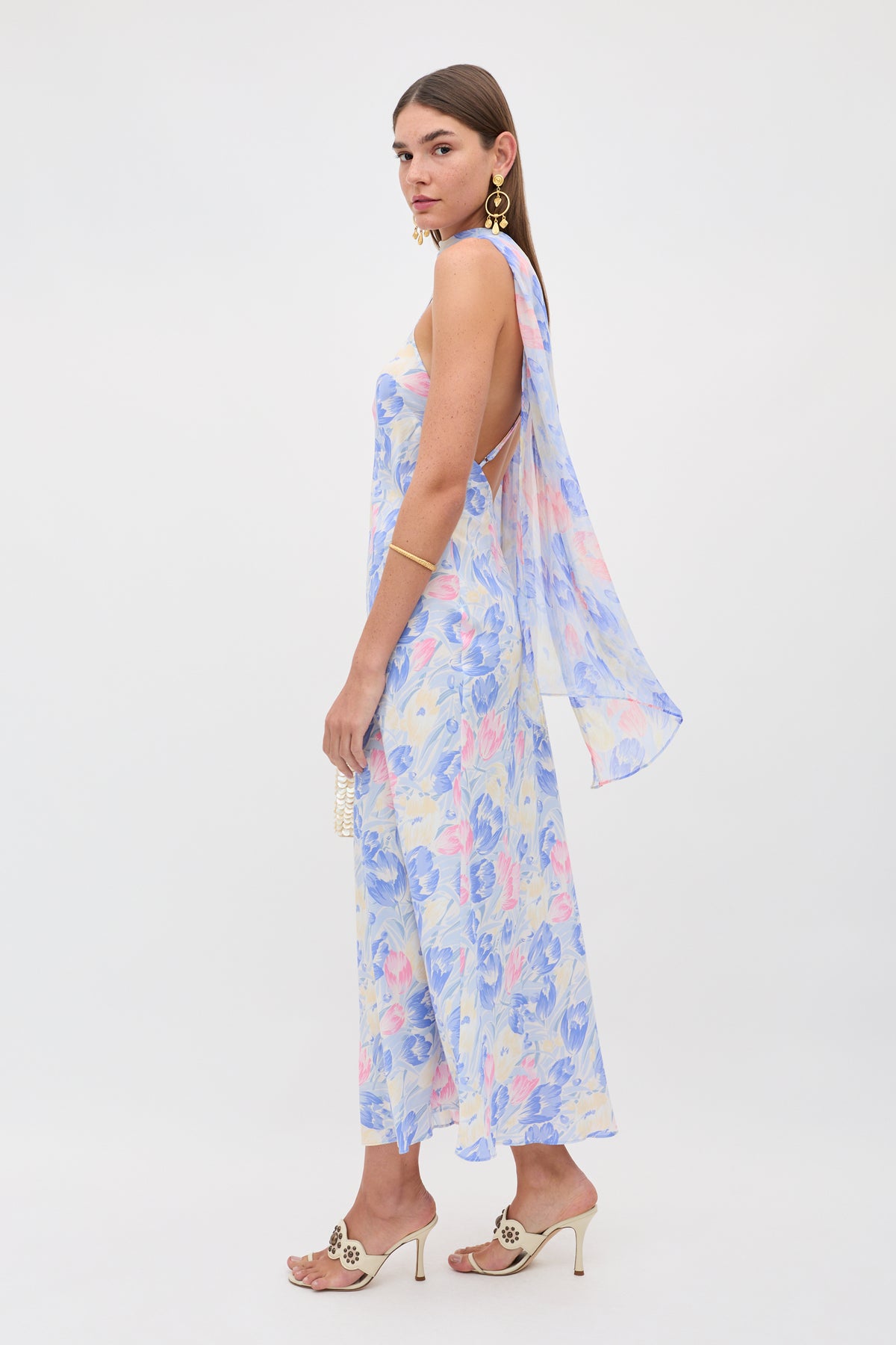 A woman stands sideways wearing the Hayley - Tulip Fields Blue—a flowy, sleeveless silk crepe de chine dress with a detachable georgette scarf—accessorized with gold jewelry, heeled sandals, and a small clutch against a plain white background.