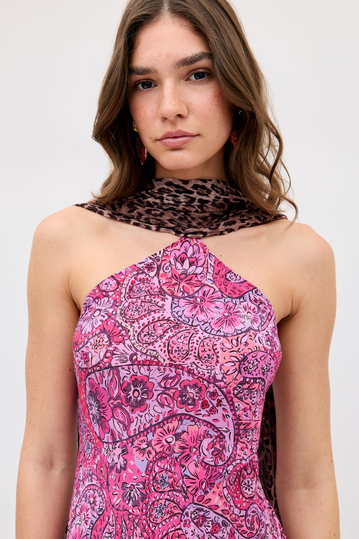 A woman with wavy brown hair wears the Hayley - Leopard Paisley Mix Multi, featuring a pink paisley one-shoulder dress and a brown leopard-print scarf, standing against a plain light background.