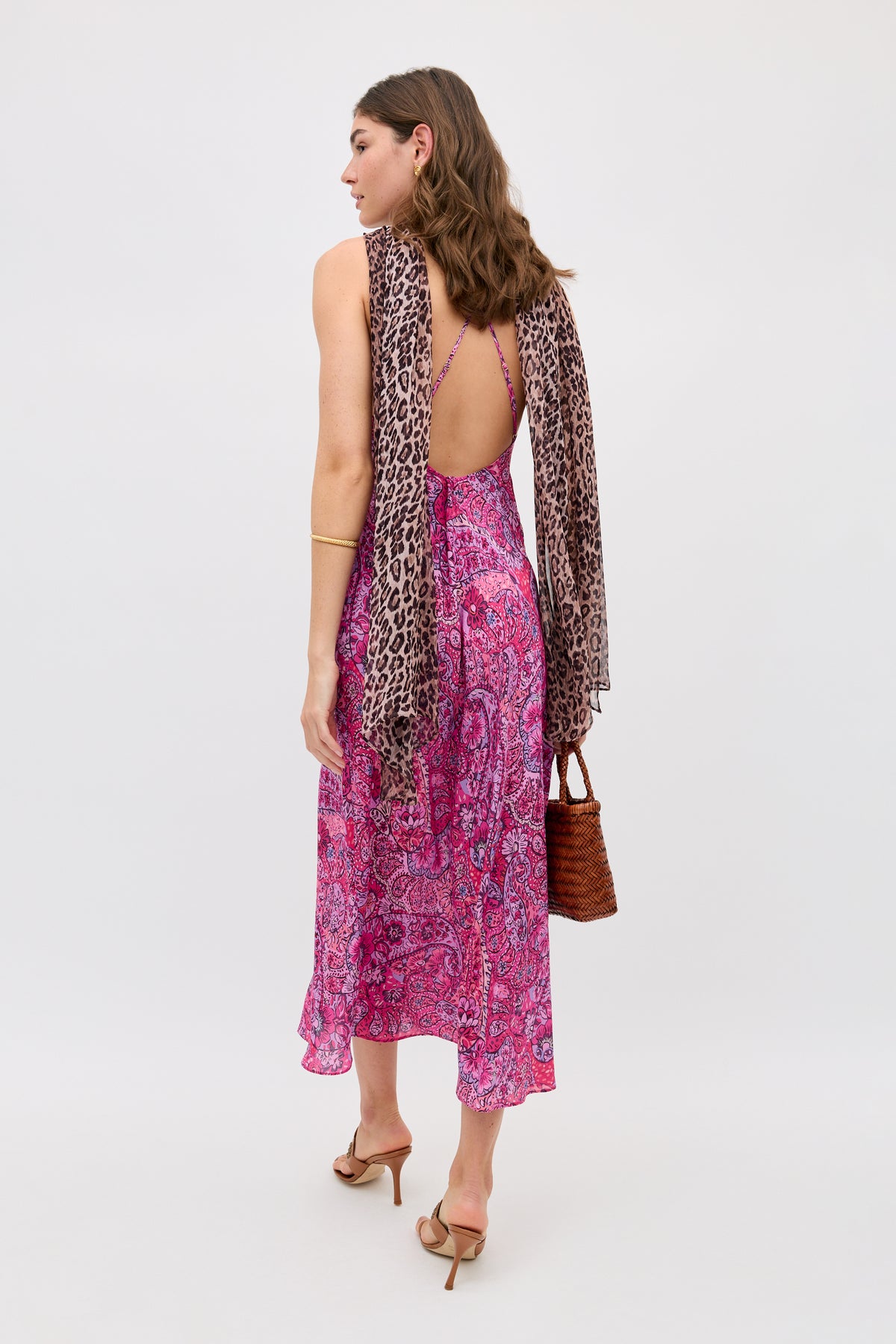 A woman with loose hair stands facing away, wearing the Hayley - Leopard Paisley Mix Multi dress with crisscross straps, tan heeled sandals, a brown woven handbag, and a leopard print scarf draped over her shoulders.