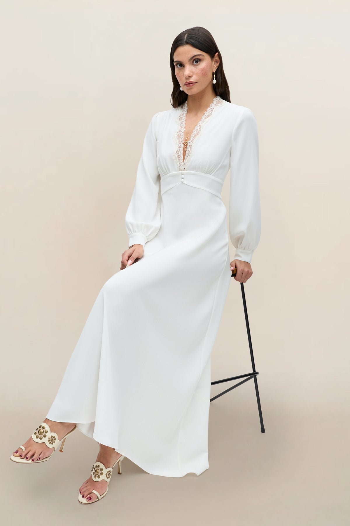 A woman wearing the Emory - Ivory Lace long-sleeved gown with delicate trim sits on a black stool against a plain beige background, paired with cream heels adorned with decorative accents.