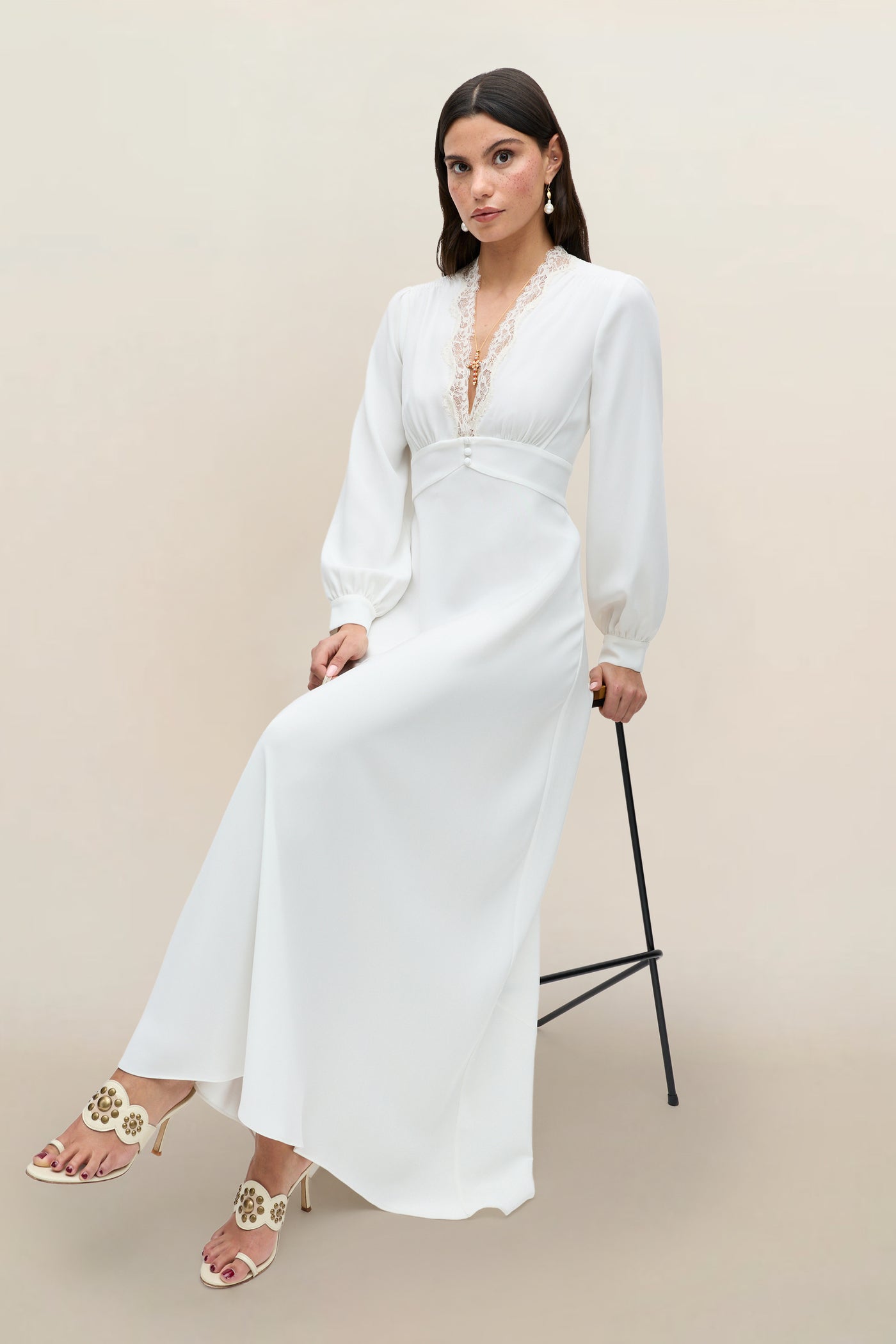 A woman wearing the Emory - Ivory Lace long-sleeved gown with delicate trim sits on a black stool against a plain beige background, paired with cream heels adorned with decorative accents.