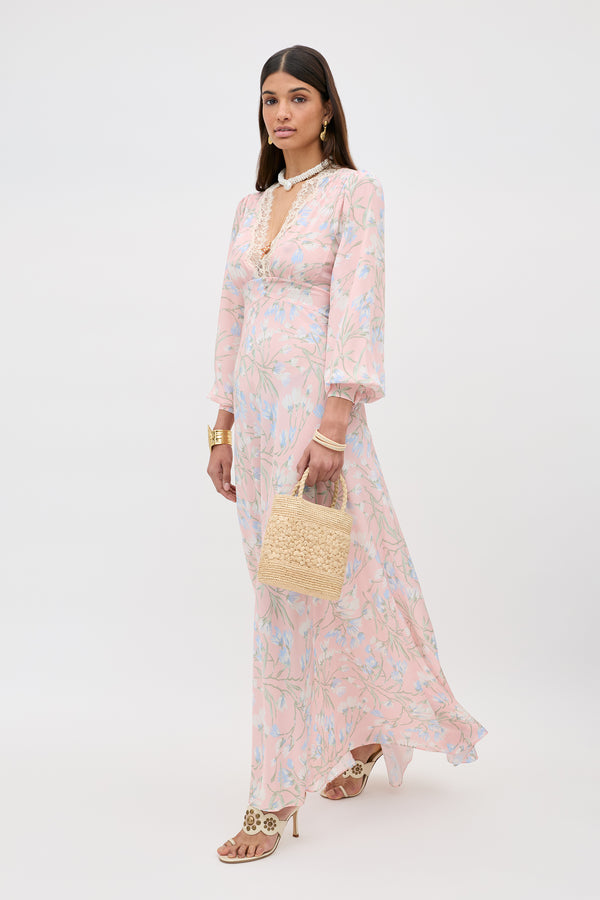 A woman models the Emory - Watercolour Floral Pink dress with long sleeves, paired with a woven handbag, gold jewelry, and sandals, standing against a plain white background.