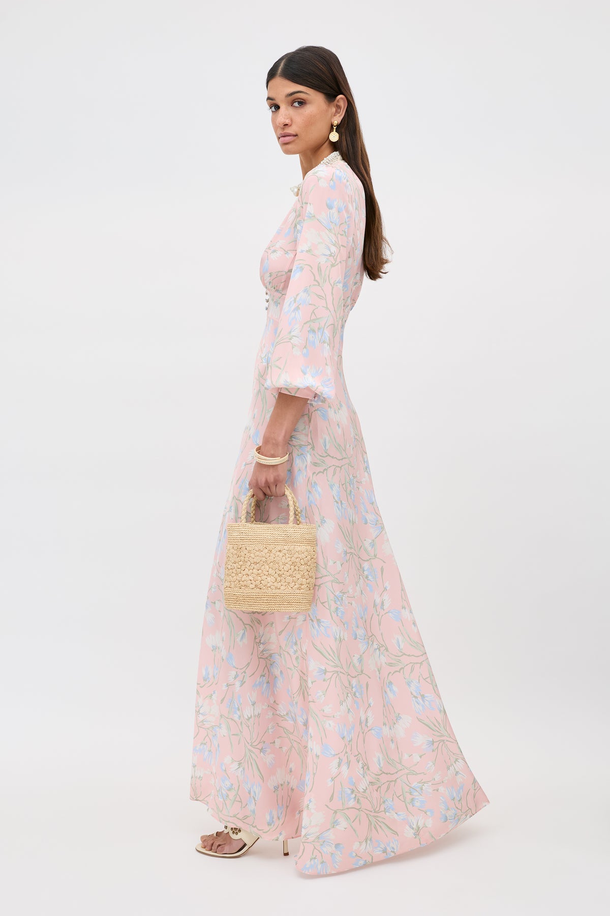 A woman in the Emory - Watercolour Floral Pink dress with bell sleeves holds a woven straw handbag. She has long brown hair, statement earrings, and stands in profile against a plain white background.