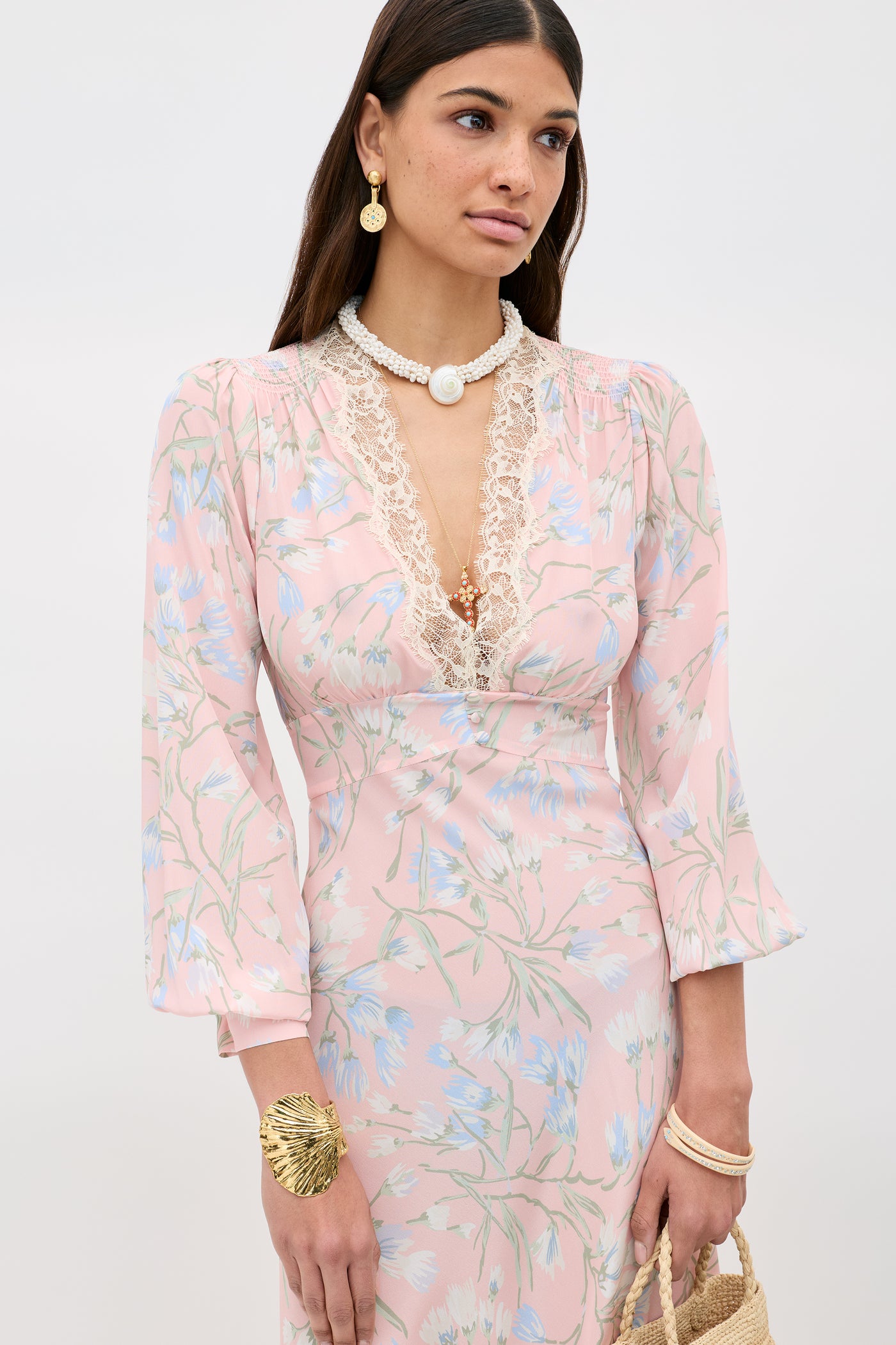 A woman in the Emory - Watercolour Floral Pink dress with lace-trimmed neckline, styled with pearl and gold jewelry—a standout gold shell bracelet—and a woven bag. She has long brown hair and a neutral expression.