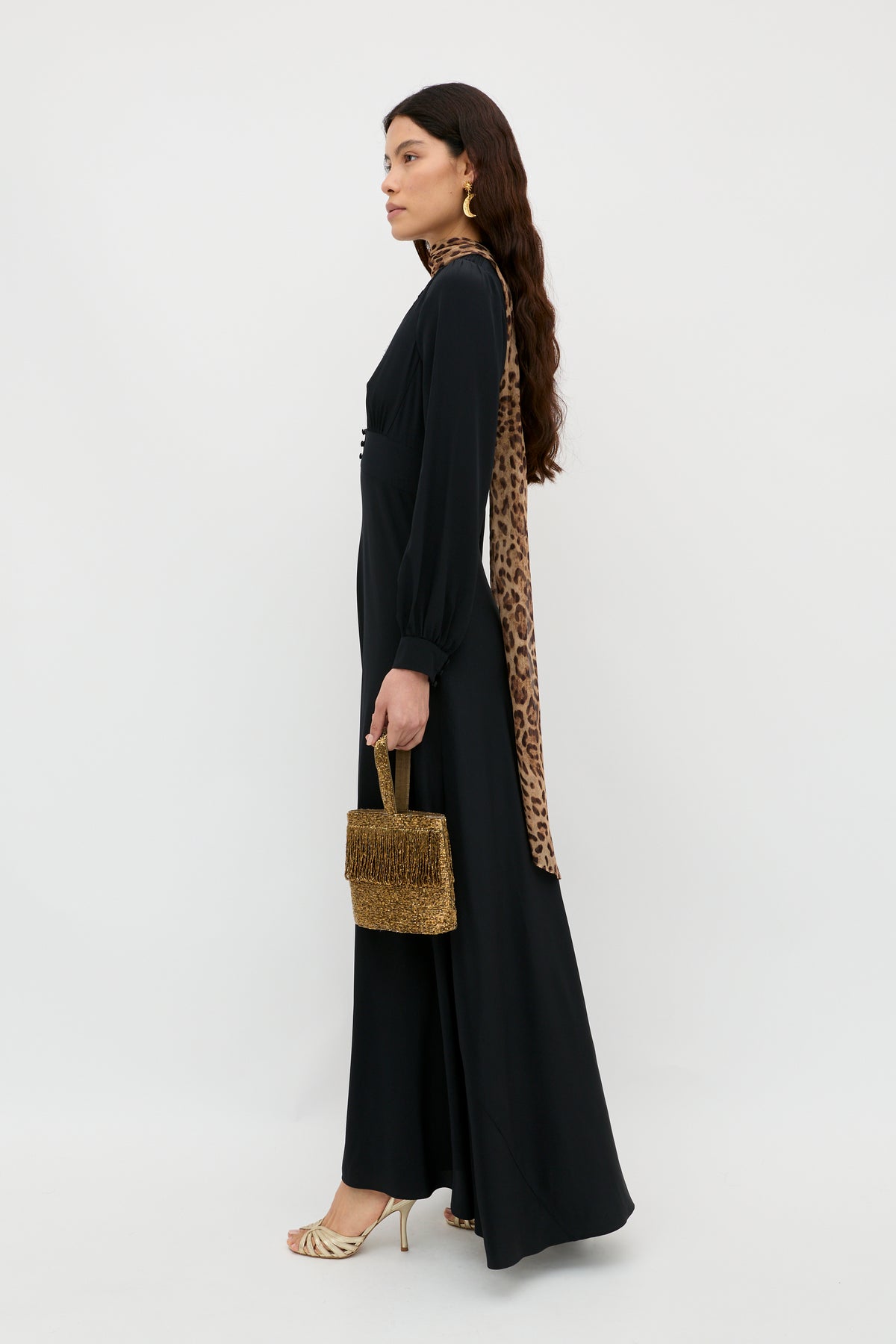 Woman in the Emory - Black dress, an elegant black silk crepe de chine piece, styled with a leopard-print scarf, gold earrings, gold heeled sandals, and a brown textured handbag. Standing sideways against a plain white background, she exudes an effortlessly chic look perfect for any special occasion.