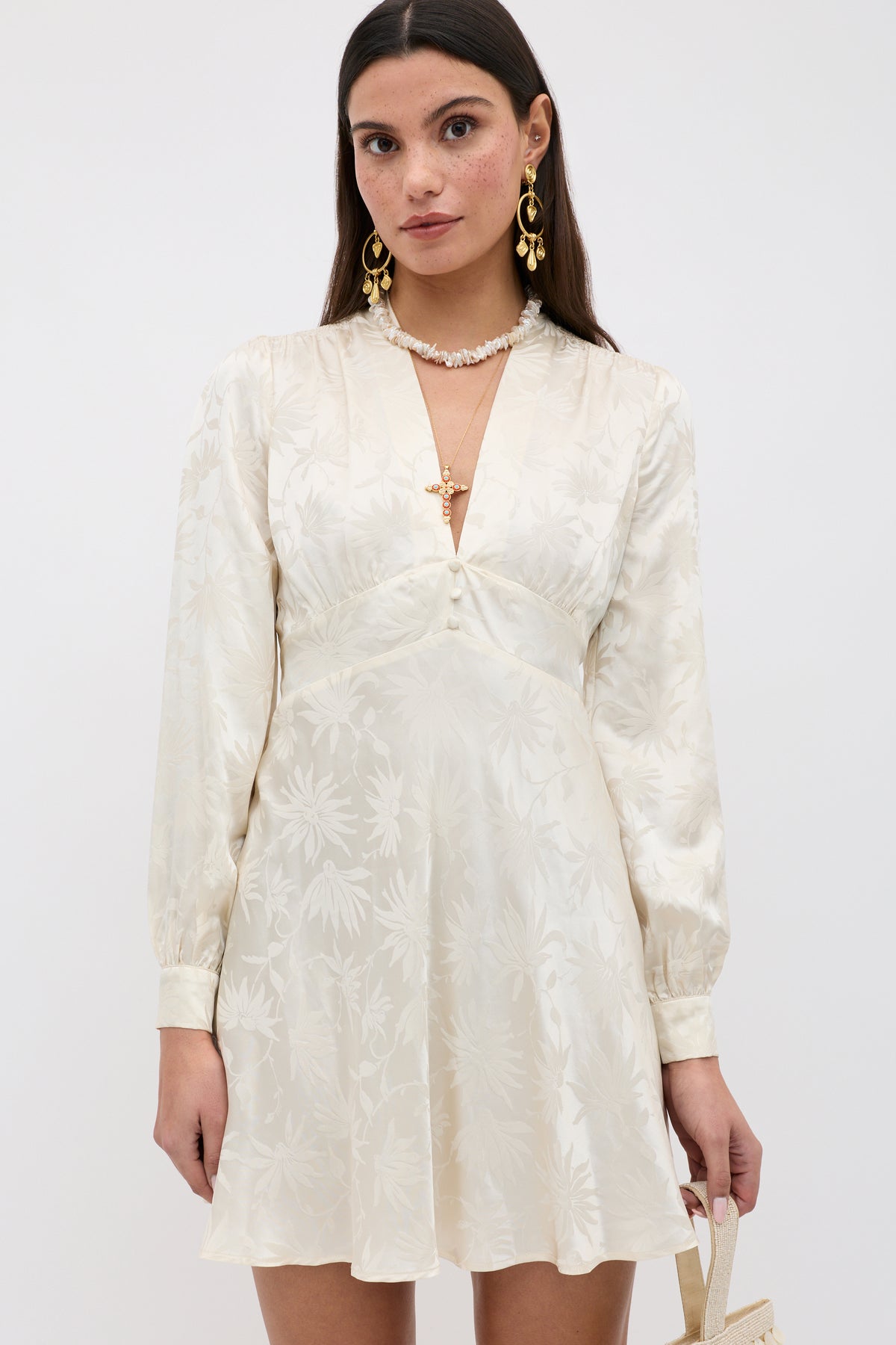 A woman with straight brown hair wears the Hebe - Daisy Jacquard Champagne mini dress, paired with gold earrings, a necklace, and holding a small handbag against a plain background.