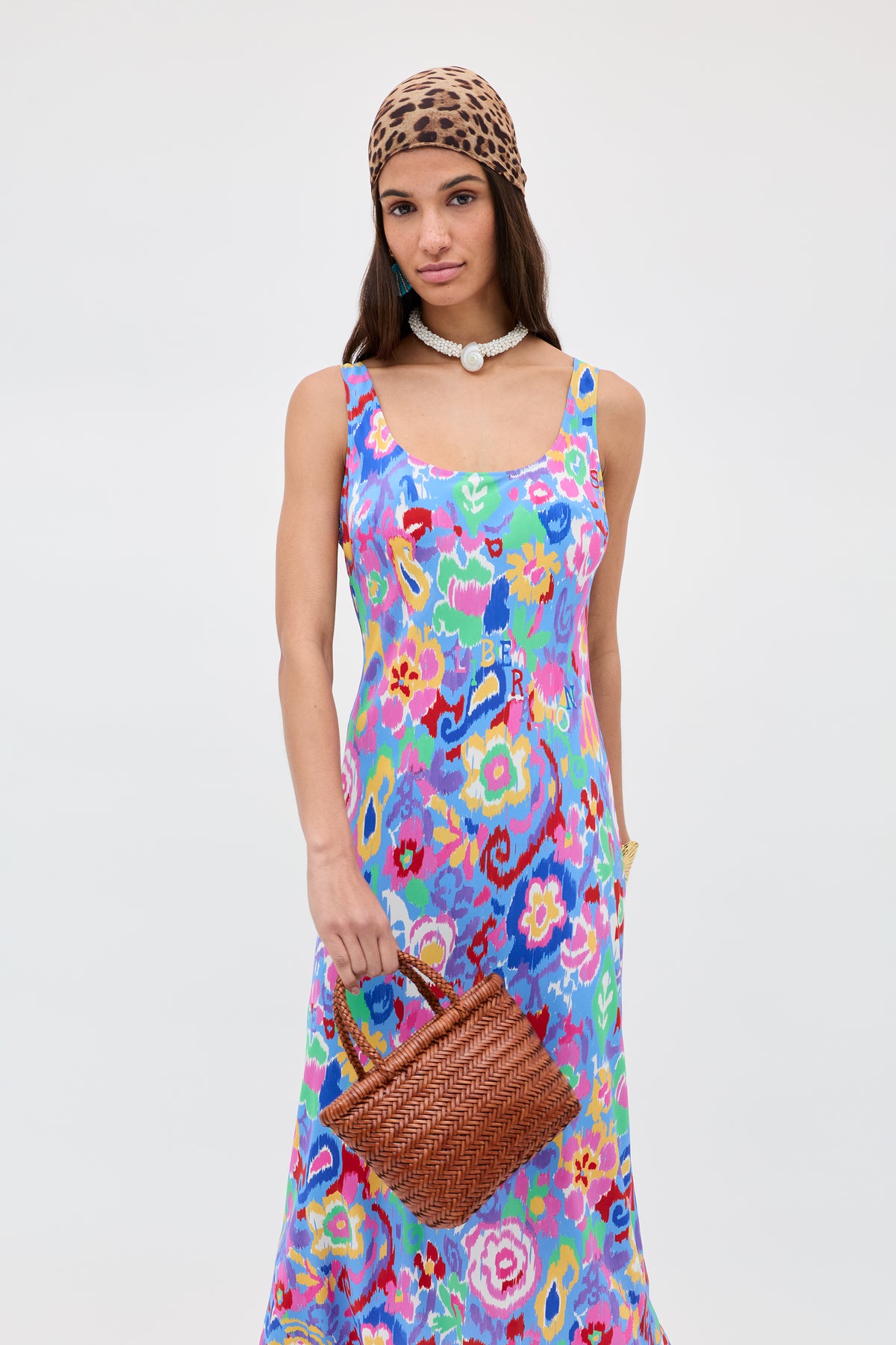 A woman in a colorful floral dress, leopard print headscarf, and white beaded necklace holds the Lysia - Tan woven bag—showcasing craftsmanship reminiscent of Dragon Diffusion—against a plain light background.