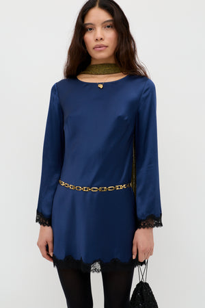 A woman in the Ridley - Midnight Navy long-sleeved satin dress with black lace trim, styled with a gold chain belt, gold scarf, and black handbag. She has long dark hair and a neutral expression.