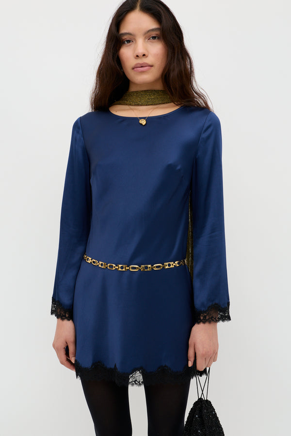 A woman in the Ridley - Midnight Navy long-sleeved satin dress with black lace trim, styled with a gold chain belt, gold scarf, and black handbag. She has long dark hair and a neutral expression.