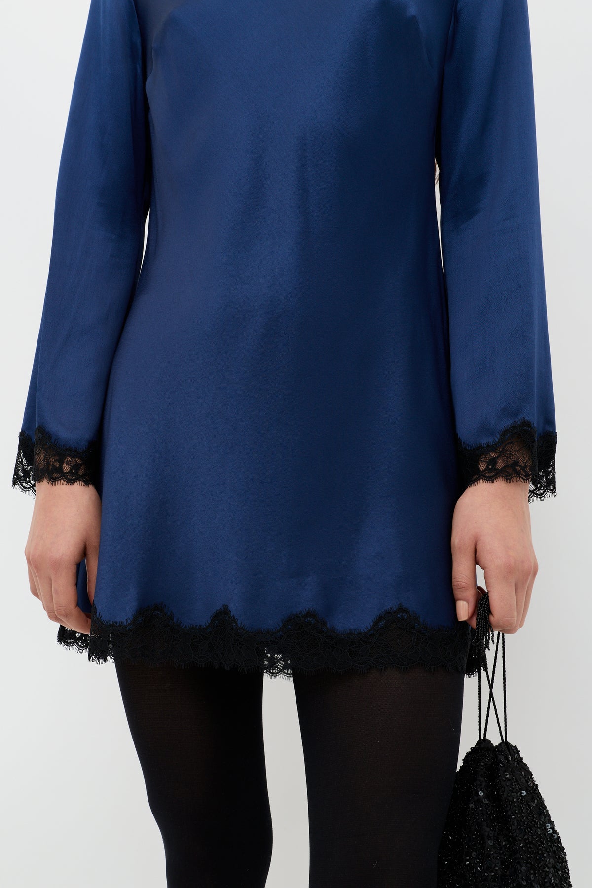 A person wearing the Ridley - Midnight Navy long-sleeve satin dress with black lace trim, paired with black tights and holding a small black beaded bag. Only the torso and upper legs are visible.