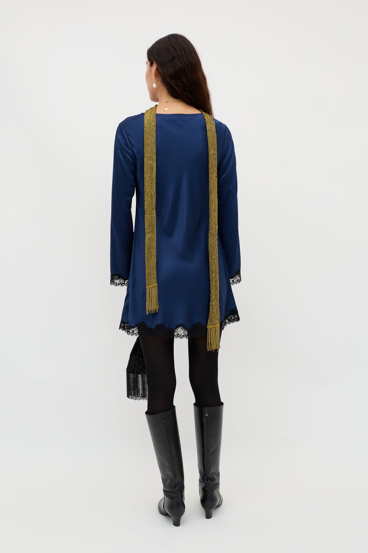 A woman faces away, wearing the Ridley - Midnight Navy long-sleeved dress with lace trim, black tights, tall black boots, a long gold scarf, and holding a small black bag against a plain white background.