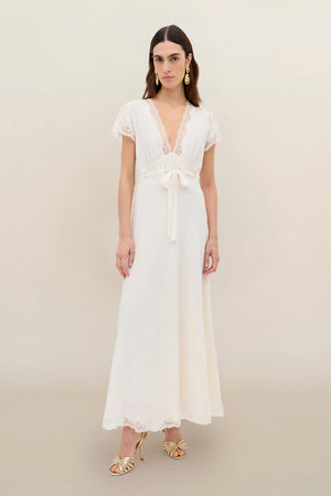 Wearing the Marlice - Pearl, a long, vintage-inspired white dress with lace trim and a deep V-neckline, she stands against a light background. Her look is finished with gold earrings, open-toe heels, and loose, center-parted hair.