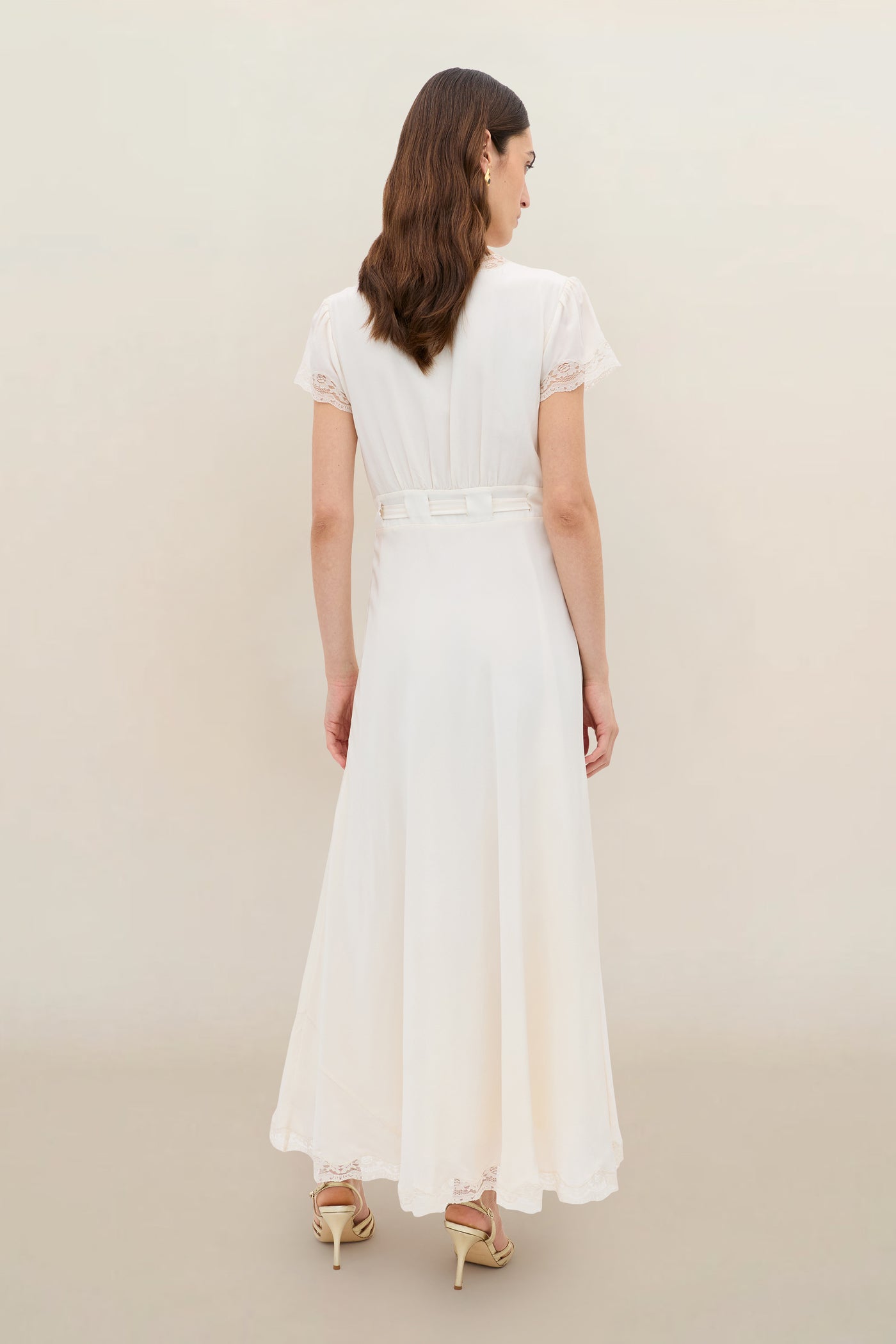 A woman with wavy brown hair wears the Marlice - Pearl: an ankle-length white dress with short sleeves, lace trim, belted waist, and satin bow. She pairs it with beige heeled sandals. She stands facing away against a plain, light background.