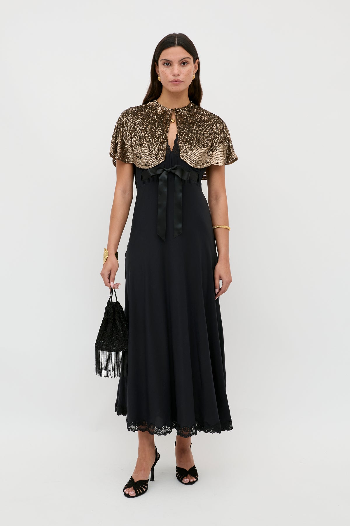 Wearing the Marlice - Black long dress with lace trim, a woman stands against a white background. She completes her look with black heels, a shiny gold capelet, a black fringe bag, gold jewelry, and sleek middle-parted hair.