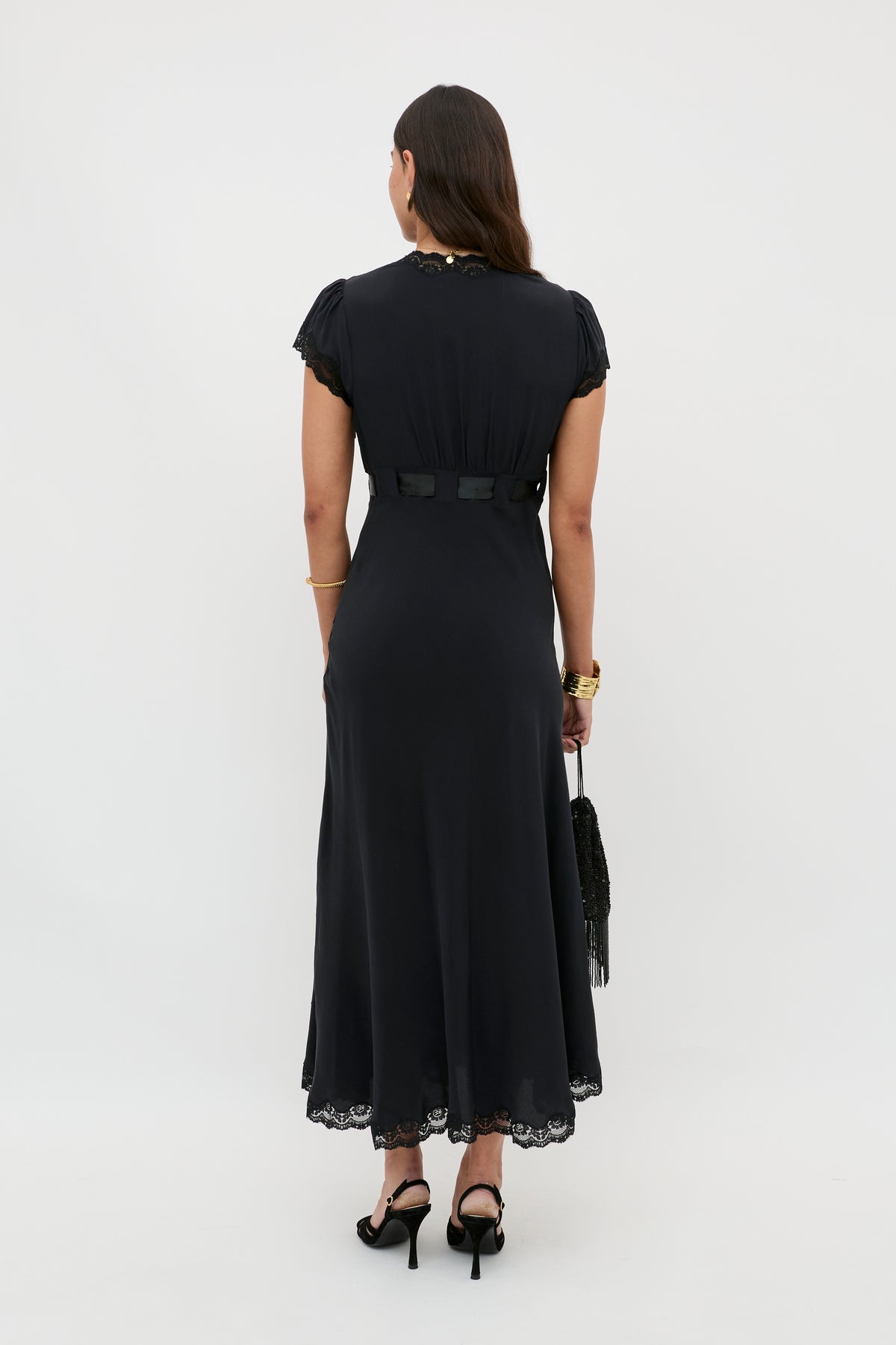 A woman stands facing away, wearing the Marlice - Black silk midi dress with lace trim. She carries a black fringe bag, wears black heels, gold bracelets, and a watch. Her dark hair is down, completing her timeless look.