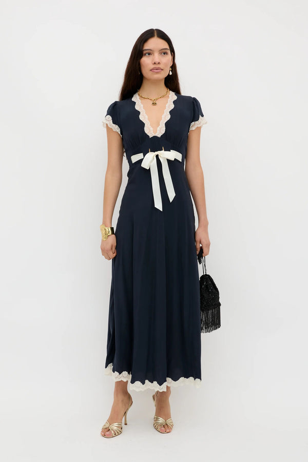 A woman with long dark hair stands against a white background wearing the Marlice - Deep Navy, an ankle-length, short-sleeved dress featuring lace trims and a cream satin bow at the waist. She accessorizes with a black bag, gold sandals, and jewelry.