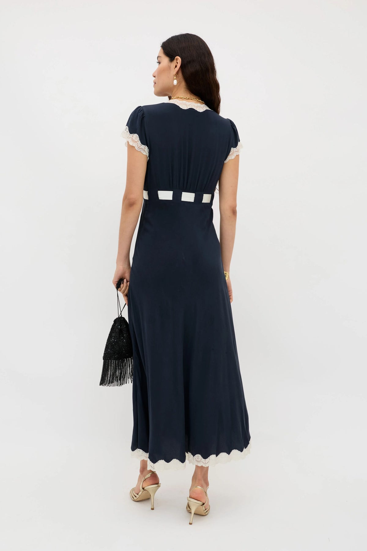 A woman stands facing away, wearing the Marlice – Deep Navy dress with white lace trim and a white square waist pattern. She carries a black fringe handbag with a satin bow, heeled sandals, and styles her long dark hair with pearl accessories.