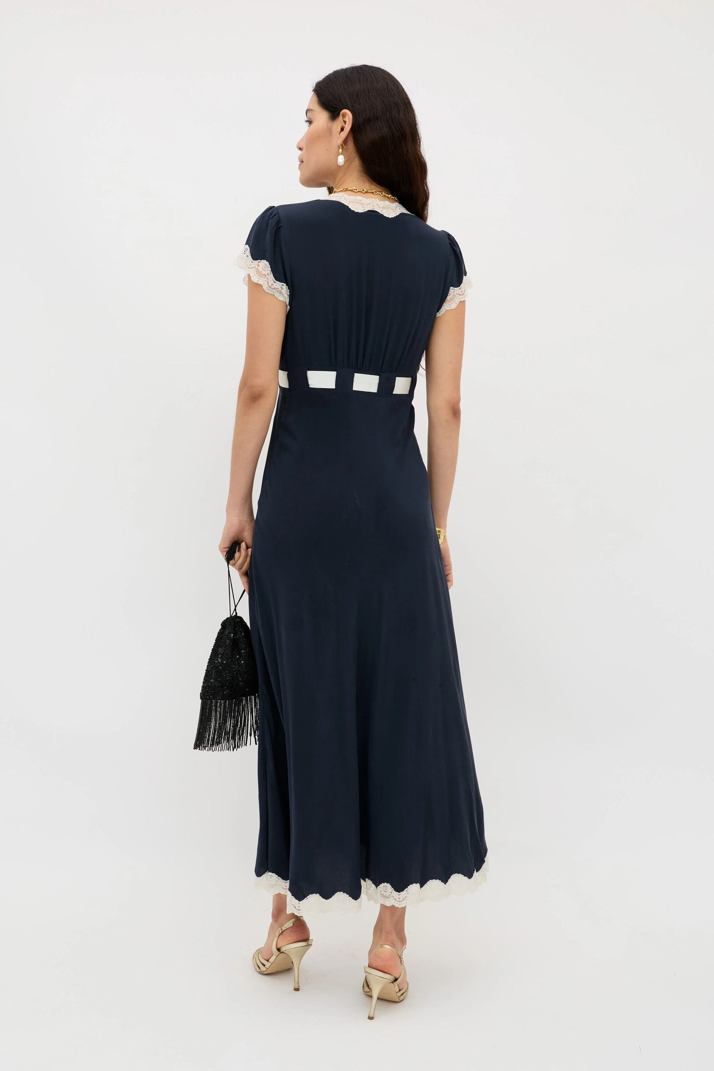 A woman stands facing away, wearing the Marlice – Deep Navy dress with white lace trim and a white square waist pattern. She carries a black fringe handbag with a satin bow, heeled sandals, and styles her long dark hair with pearl accessories.