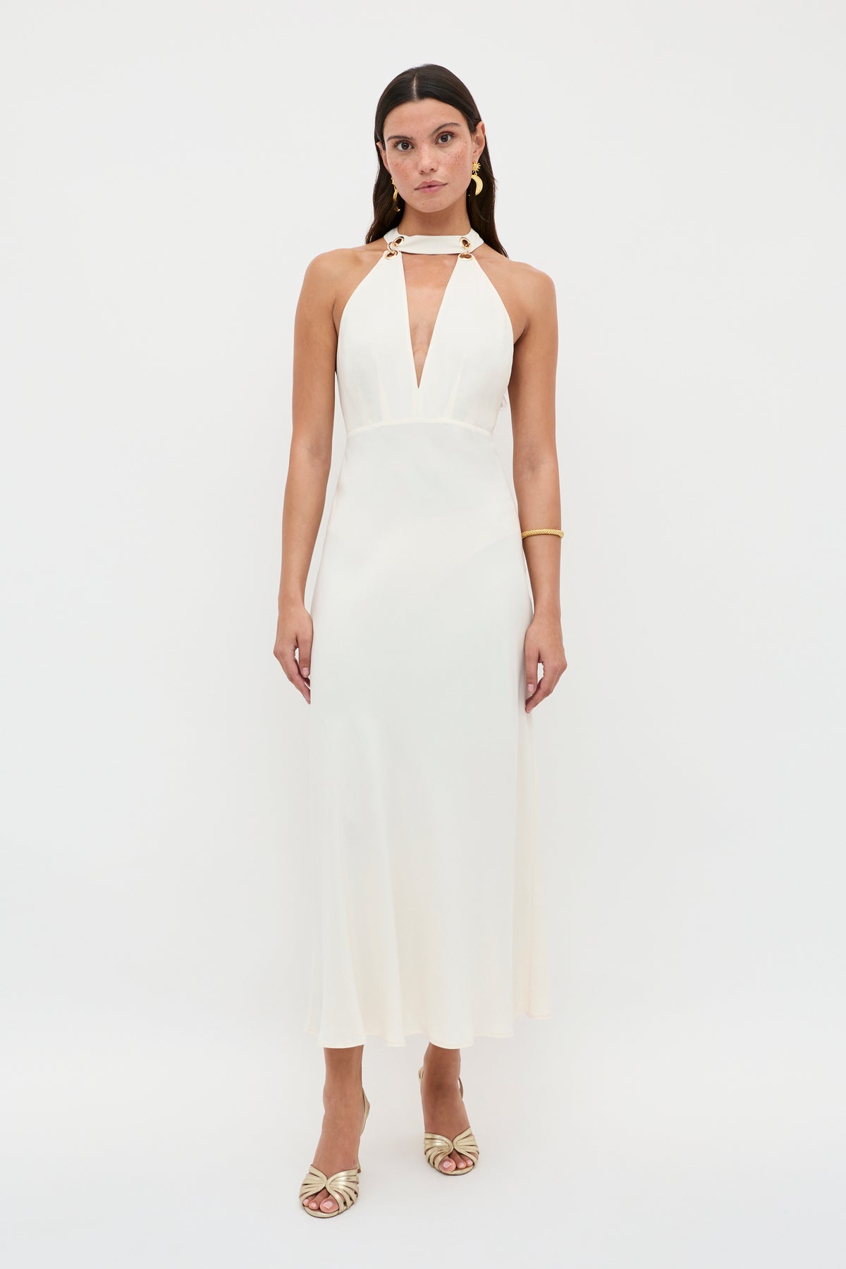 A woman stands against a white background wearing the Eleanora - Pearl dress by RIXO, featuring a sleeveless halter neck, deep V-neckline, and bias-cut hem. She accessorizes with gold earrings, a bracelet, and gold open-toe heels for a vintage-inspired look.