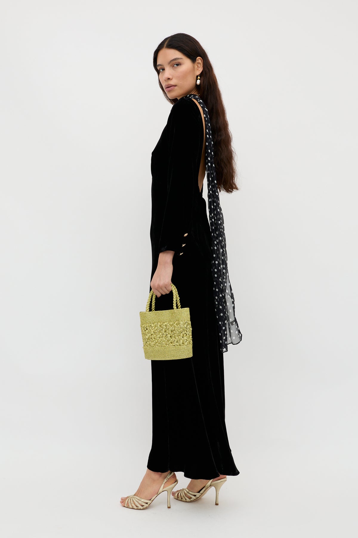 Woman in the Devina - Black elegant evening dress with long sleeves and a polka dot scarf detail, holding a yellow woven handbag, wearing gold strappy heels, against a plain white background.