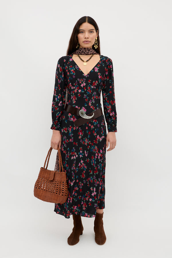 A woman poses against a white background wearing the Mavis - City Floral Black long-sleeve dress, brown ankle boots, a chunky belt with a large silver buckle, gold jewelry, and holding a woven brown handbag.